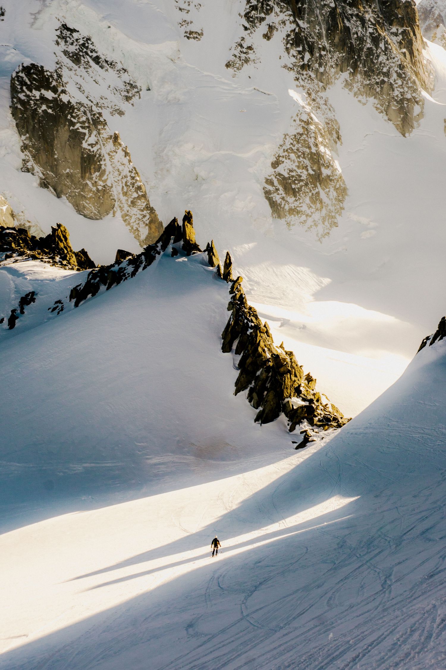 A lone skier descends a vast snow-covered alpine slope surrounded by dramatic rocky peaks bathed in golden sunlight.