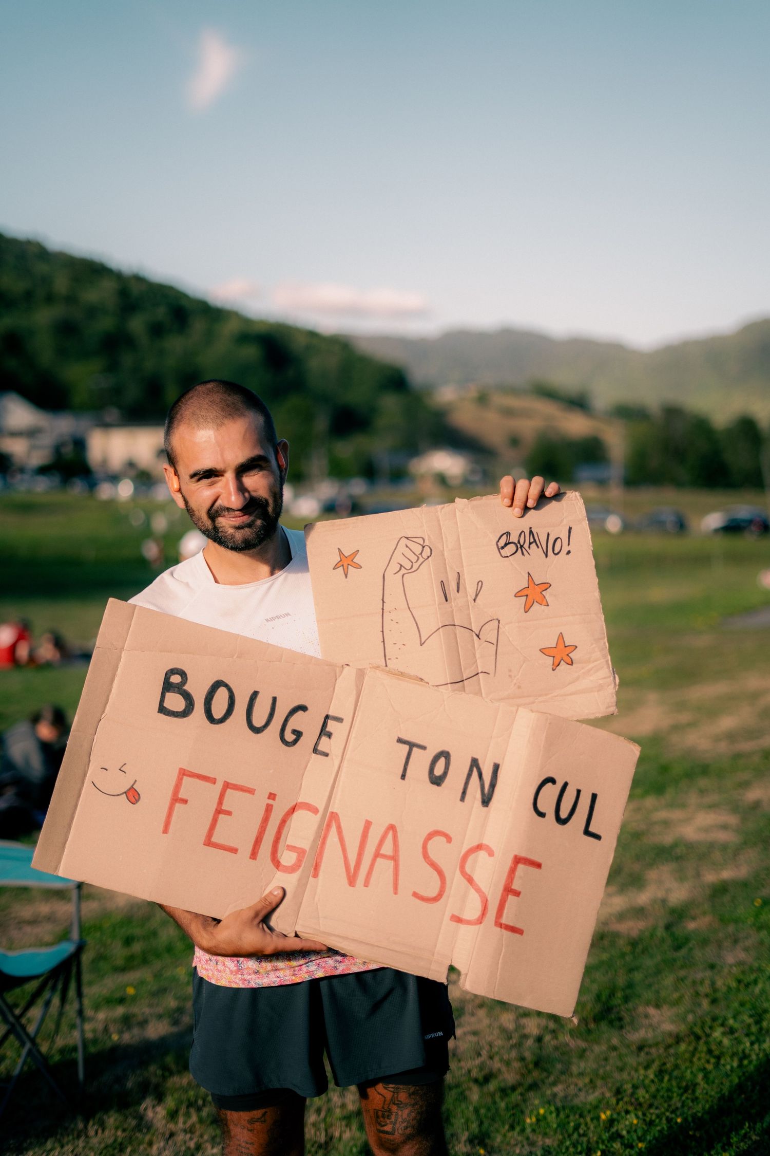 Smiling man holding cardboard signs reading Bouge Ton Cul Feignasse at outdoor sporting event.