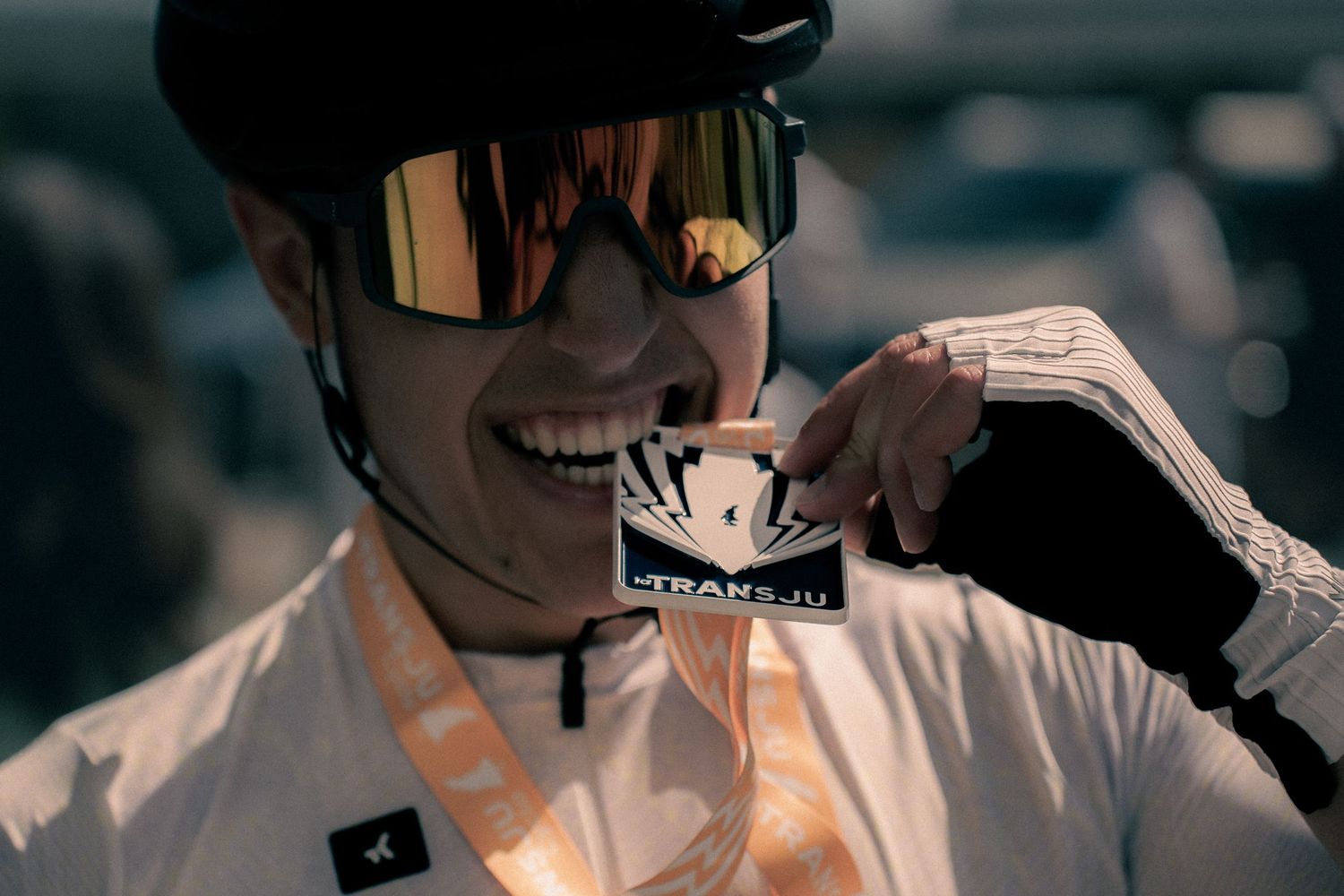 Cyclist smiling and biting a Trans-Ju race medal, wearing helmet and sunglasses.
