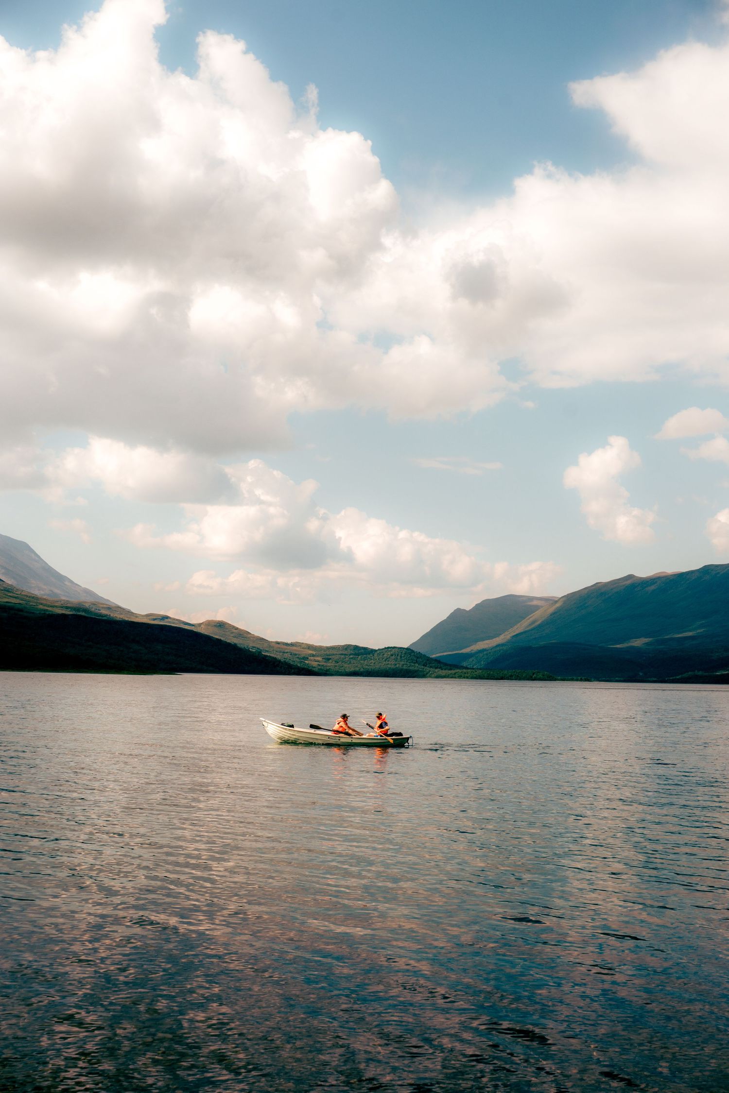 Two people rowing a white boat on a calm mountain lake under a cloudy sky.