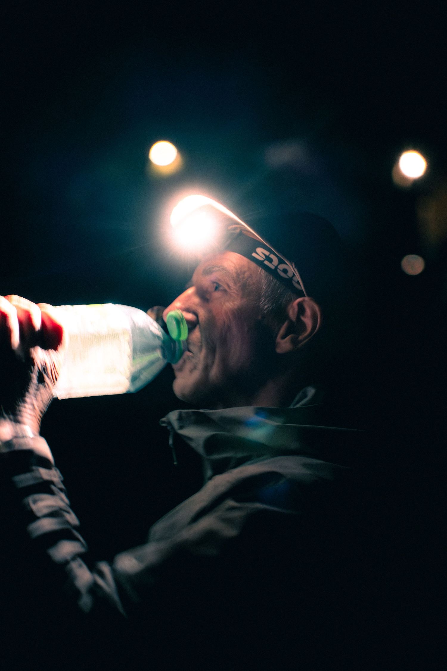 Elderly trail runner drinking from a water bottle at night with headlamp illuminating the scene.