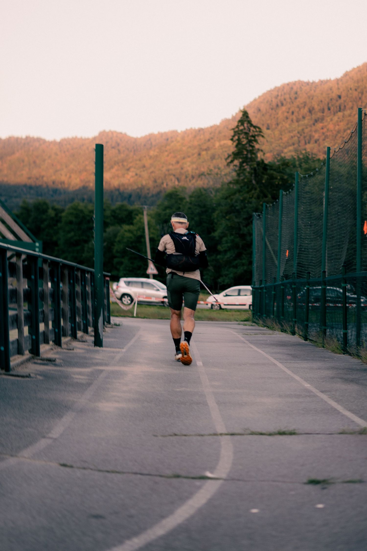 Male trail runner with hydration vest and trekking poles running along a fenced path near mountains.