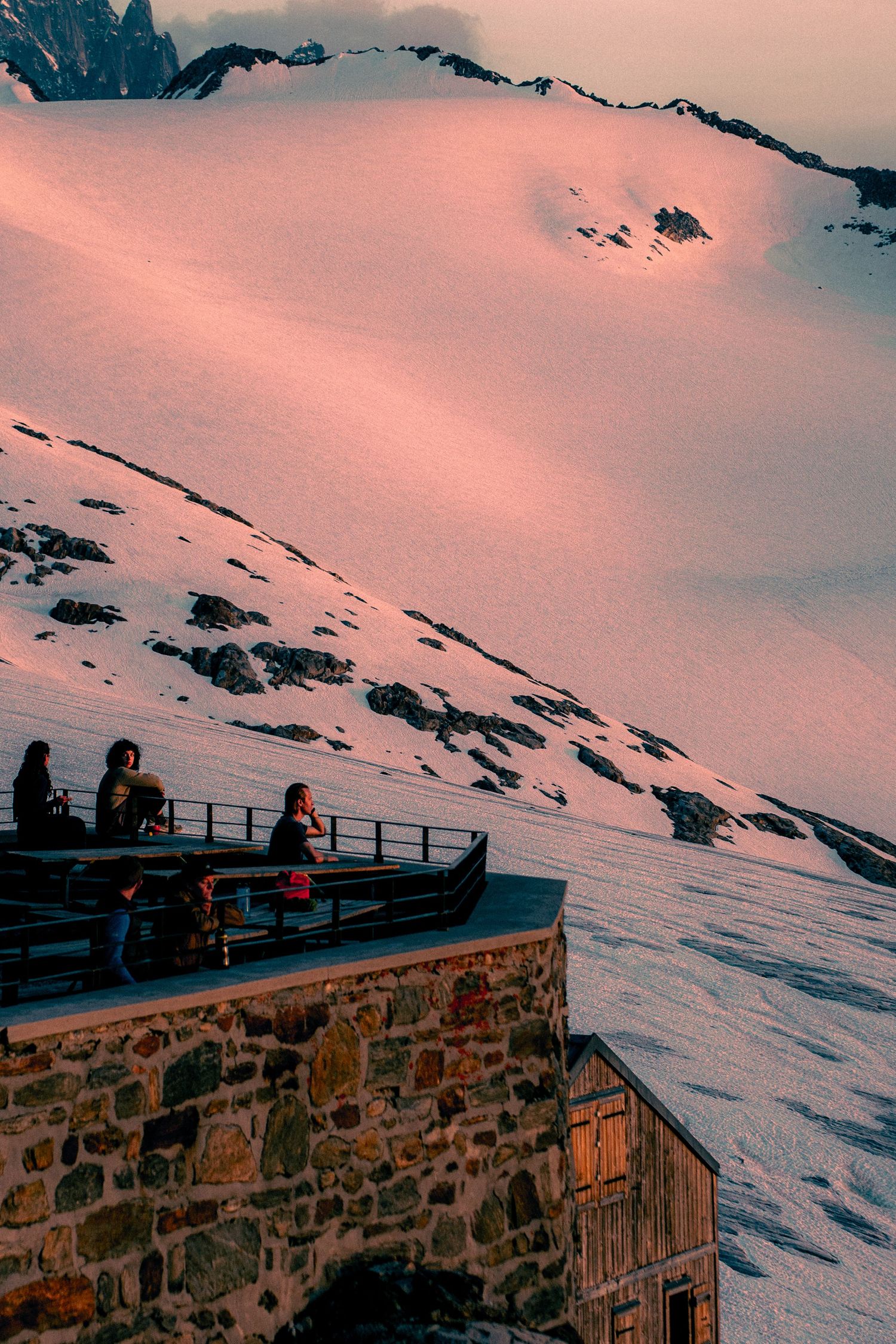 People on a stone mountain refuge terrace overlooking a vast pink-lit glacier at sunset.