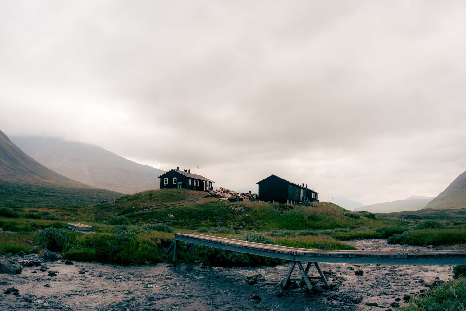 Remote dark wooden mountain cabins on a hill beside a stream in a misty Nordic valley landscape.