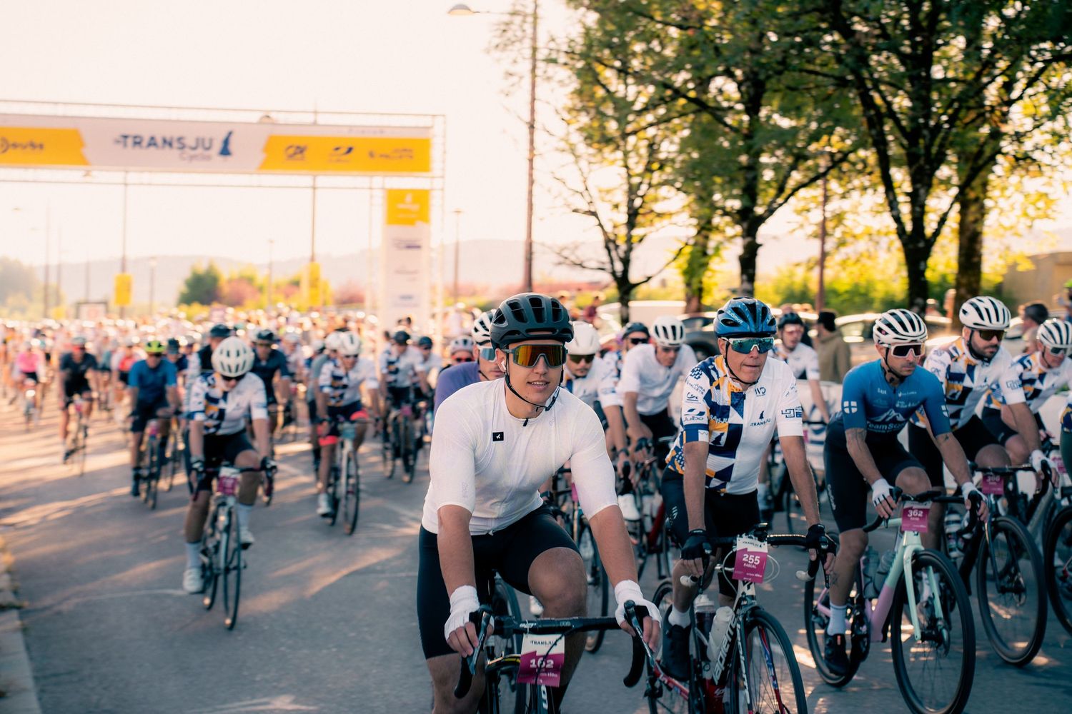 Cyclists gather at the start line of a Trans bike race event, wearing helmets and jerseys on a sunny day.
