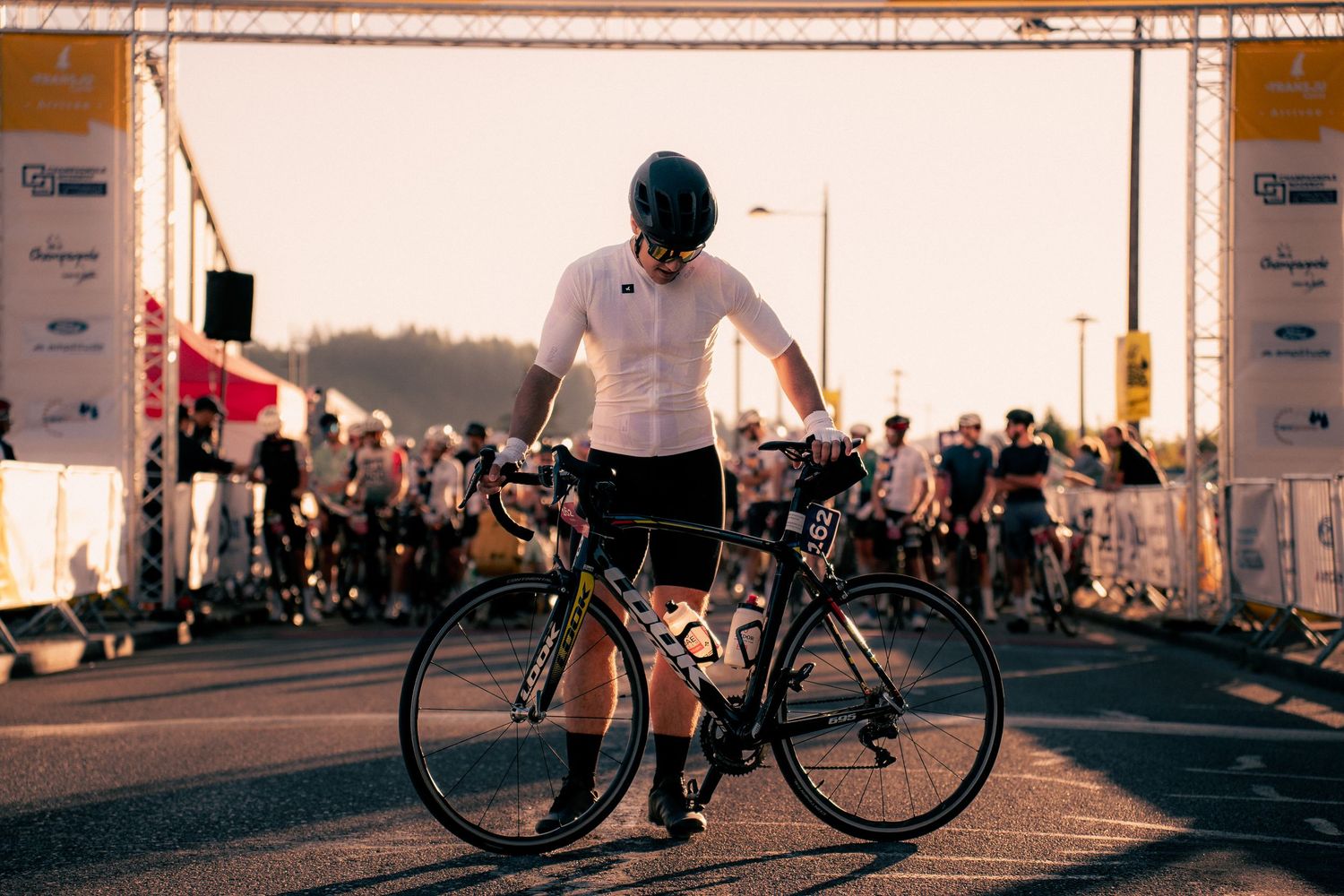 A cyclist in white jersey and helmet stands with road bike at starting line during a golden hour cycling event.