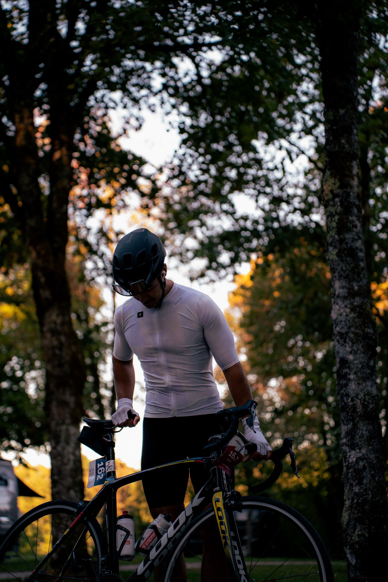 Cyclist in white jersey and helmet stands with road bike among trees in soft evening light.