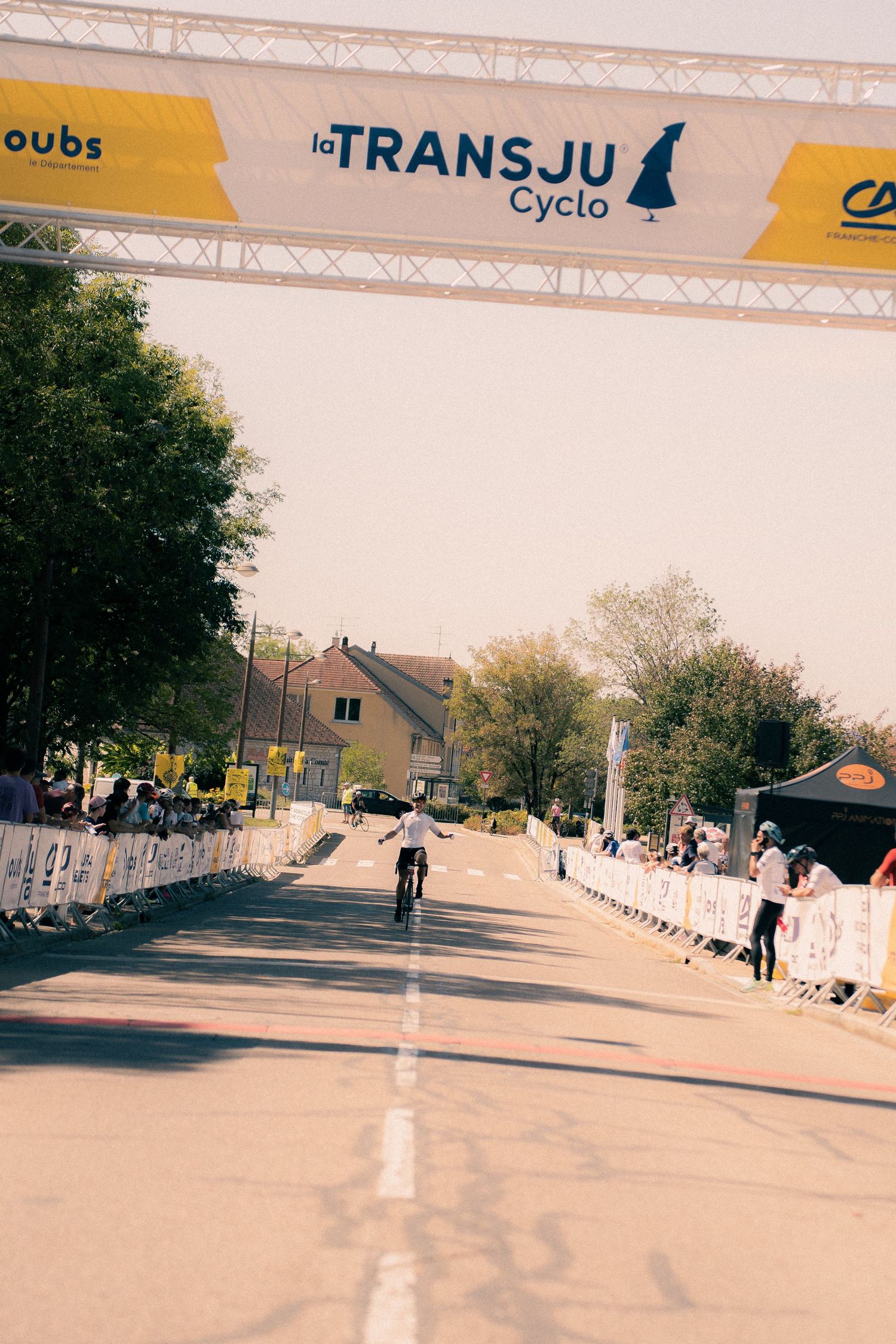 A cyclist crosses the finish line at La TransJu Cyclo race under a yellow banner on a sunny day.