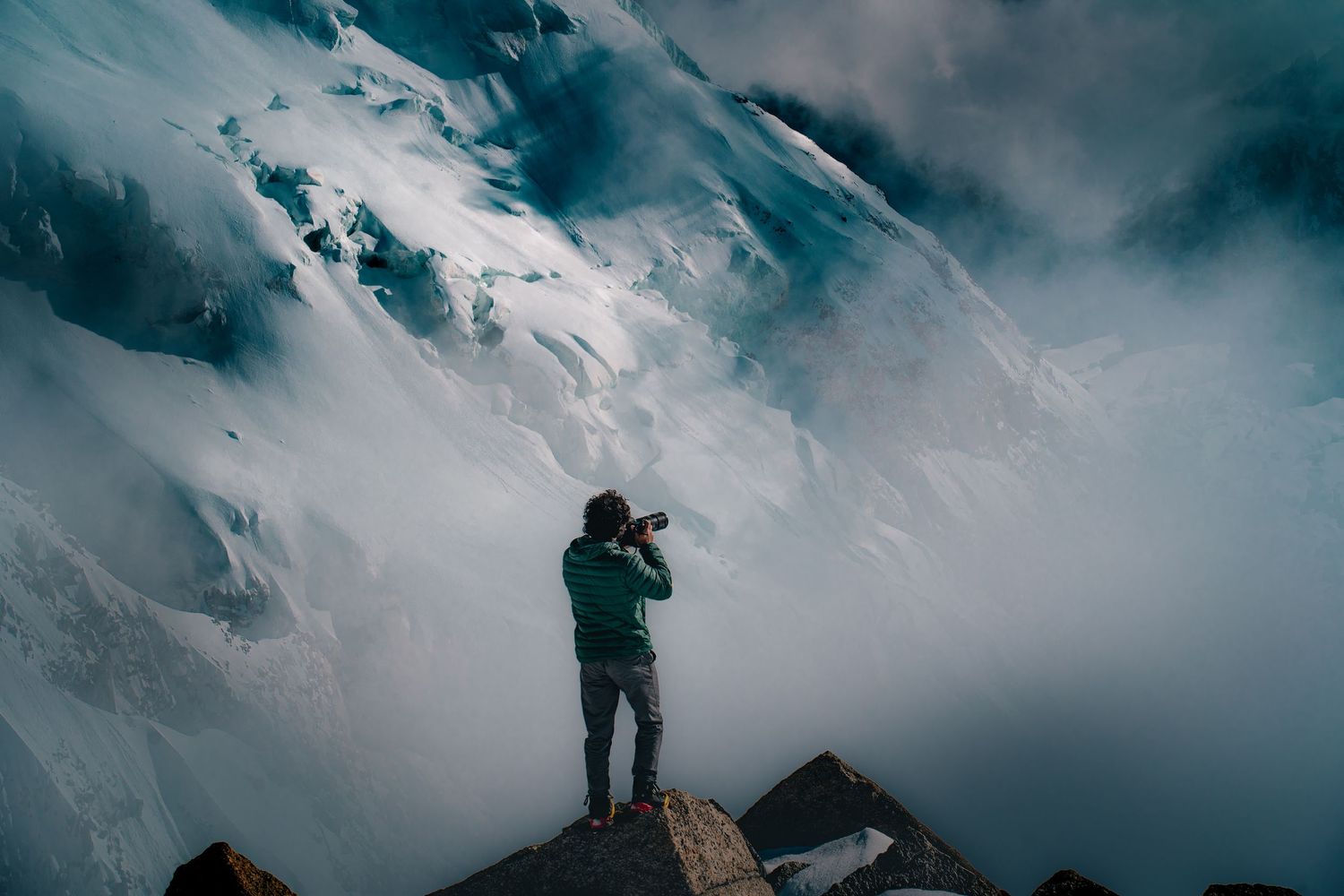 A lone photographer stands on rocky summit capturing misty snow-covered mountain peaks shrouded in clouds.