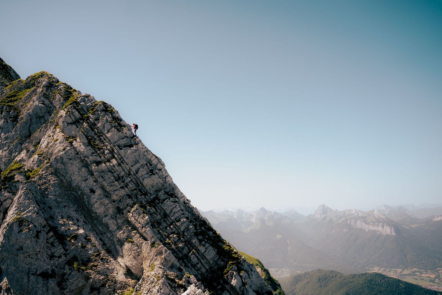 A lone climber scales a steep rocky mountain ridge under a clear blue sky with distant alpine peaks in the background.