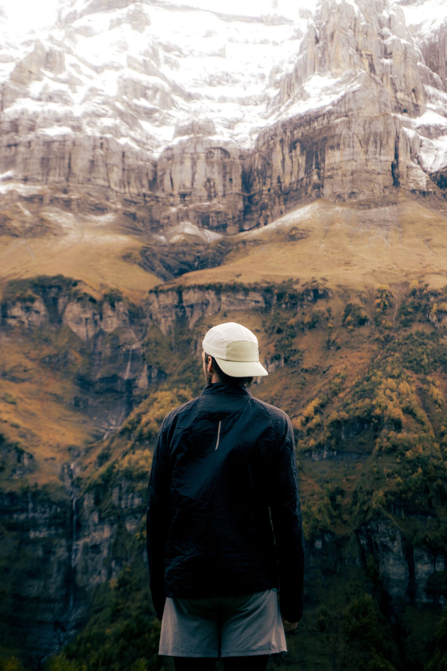 A hiker in a white cap and black jacket stands before a snow-capped rocky mountain.