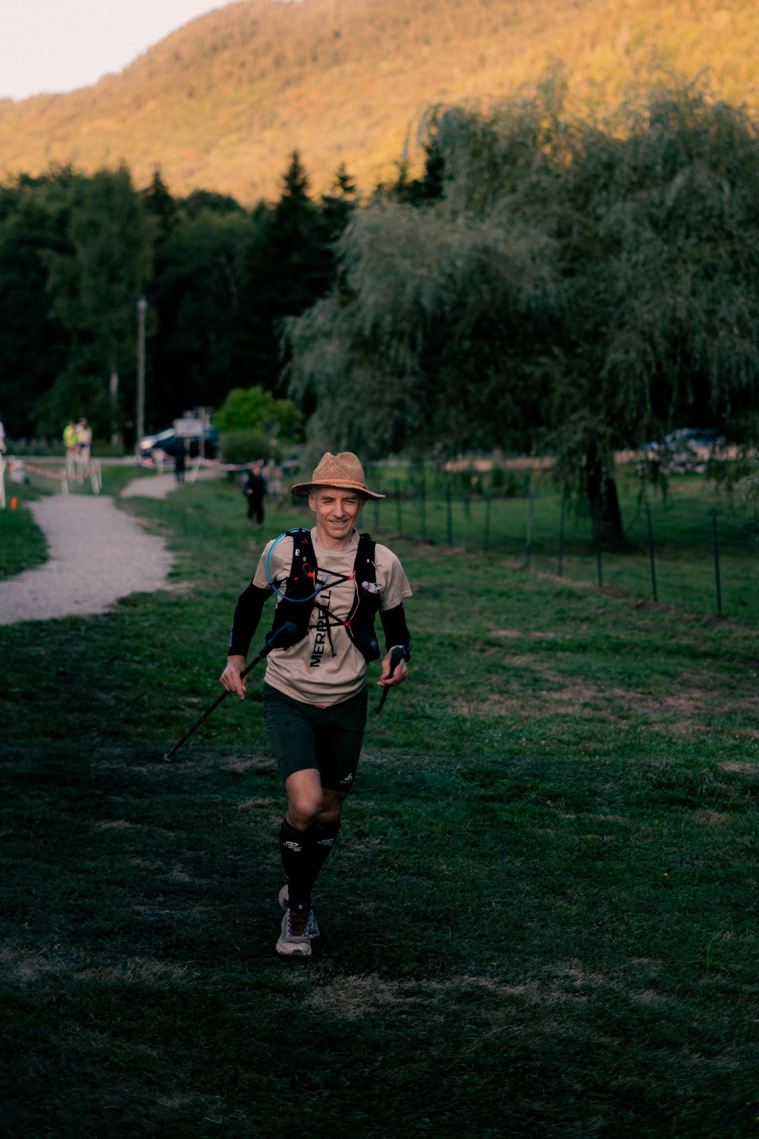 Smiling trail runner in straw hat and backpack running through park at dusk during race event.
