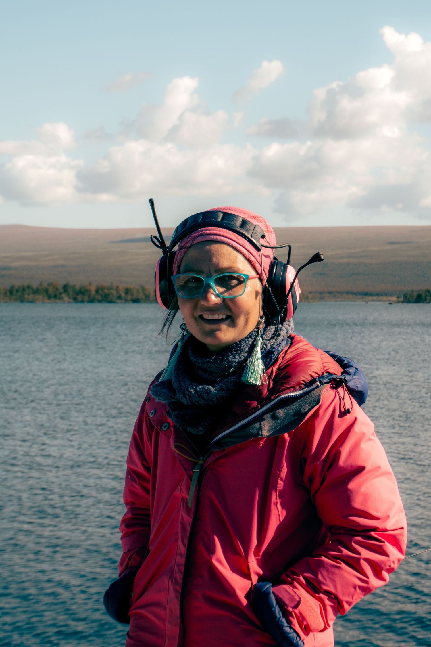 Smiling hiker in red jacket and pink hat standing by a mountain lake on a cloudy day.