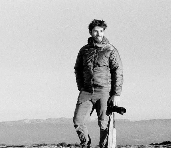 Black and white photo of a man in a puffer jacket holding a camera, standing on a rocky hilltop with mountains behind.