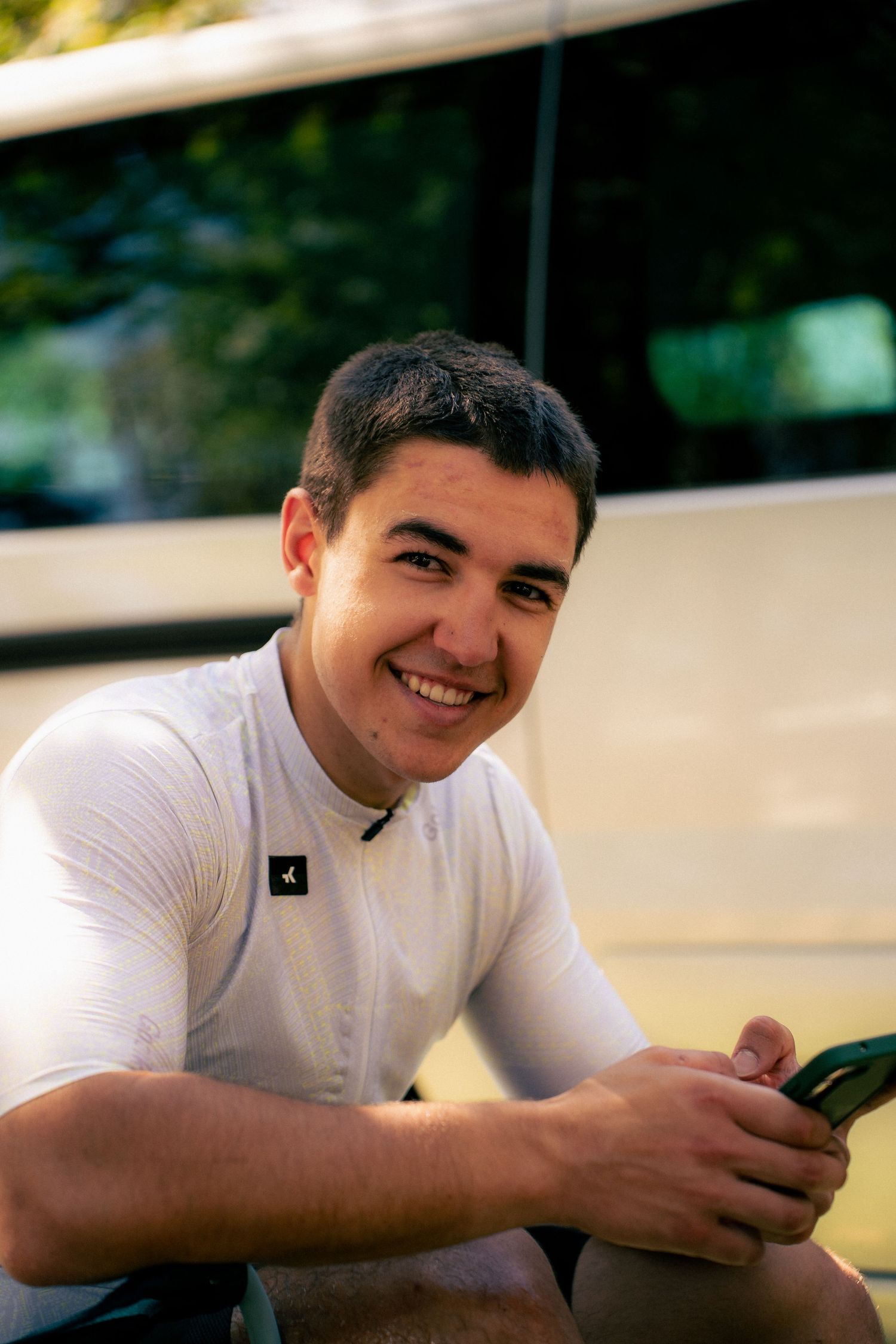 Smiling young man in white shirt sitting outdoors holding a phone, with a white vehicle and green trees in background.