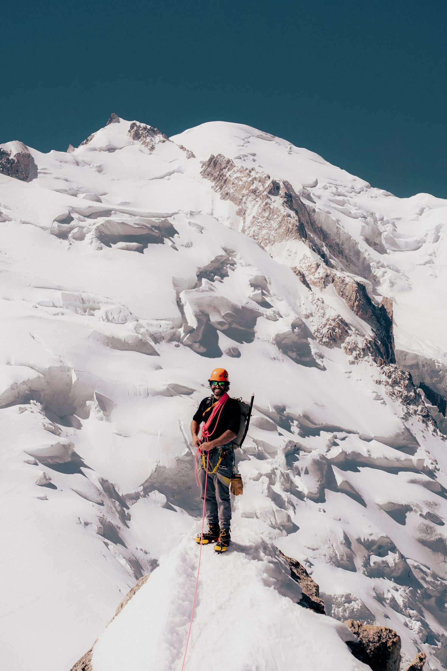 A mountaineer in helmet and harness stands on a snowy ridge with a massive glaciated peak behind under clear blue sky.