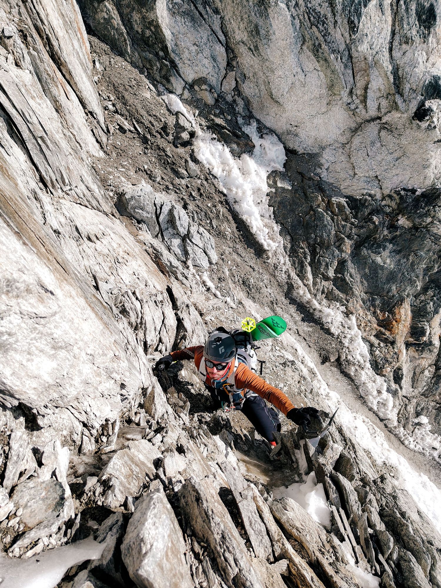 A rock climber in orange pants and green helmet scales a steep rocky mountain face with snow visible below.