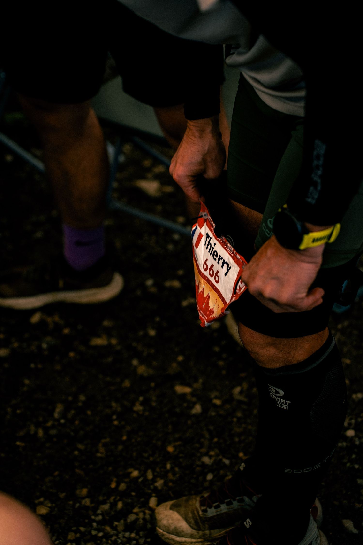 Runner attaching a race bib numbered 566 with the name Thierry to their leg in darkness.