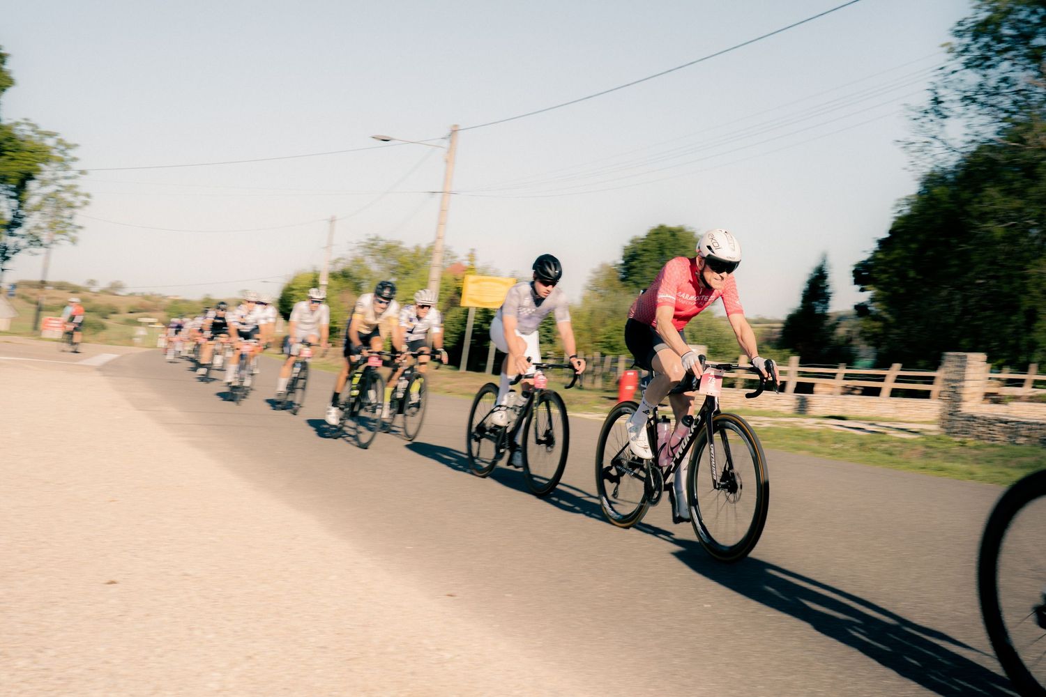 Group of cyclists racing on a sunny rural road, wearing colorful jerseys and helmets during a cycling event.