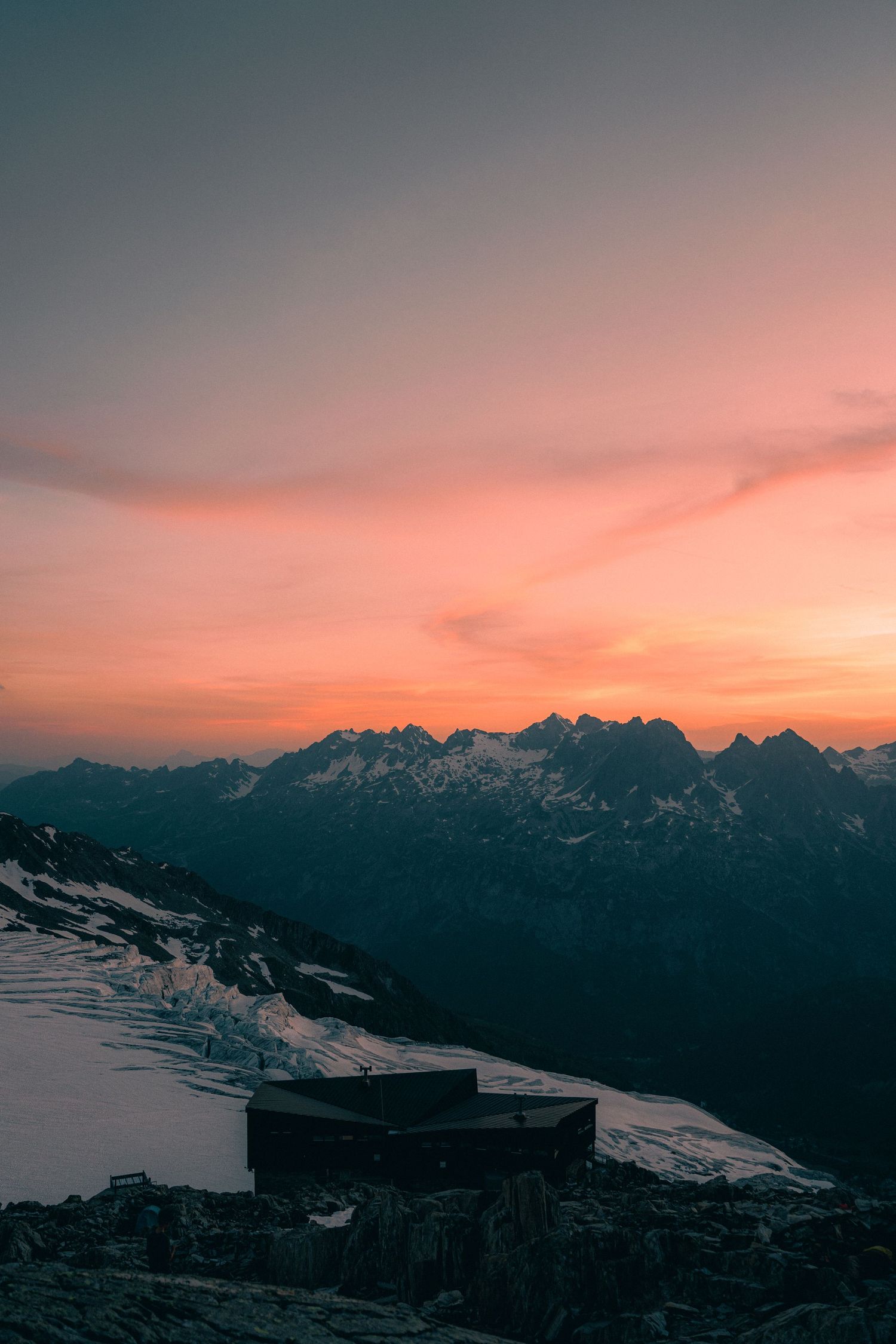 Mountain refuge silhouetted against a vivid orange and pink sunset sky over snowy alpine peaks.