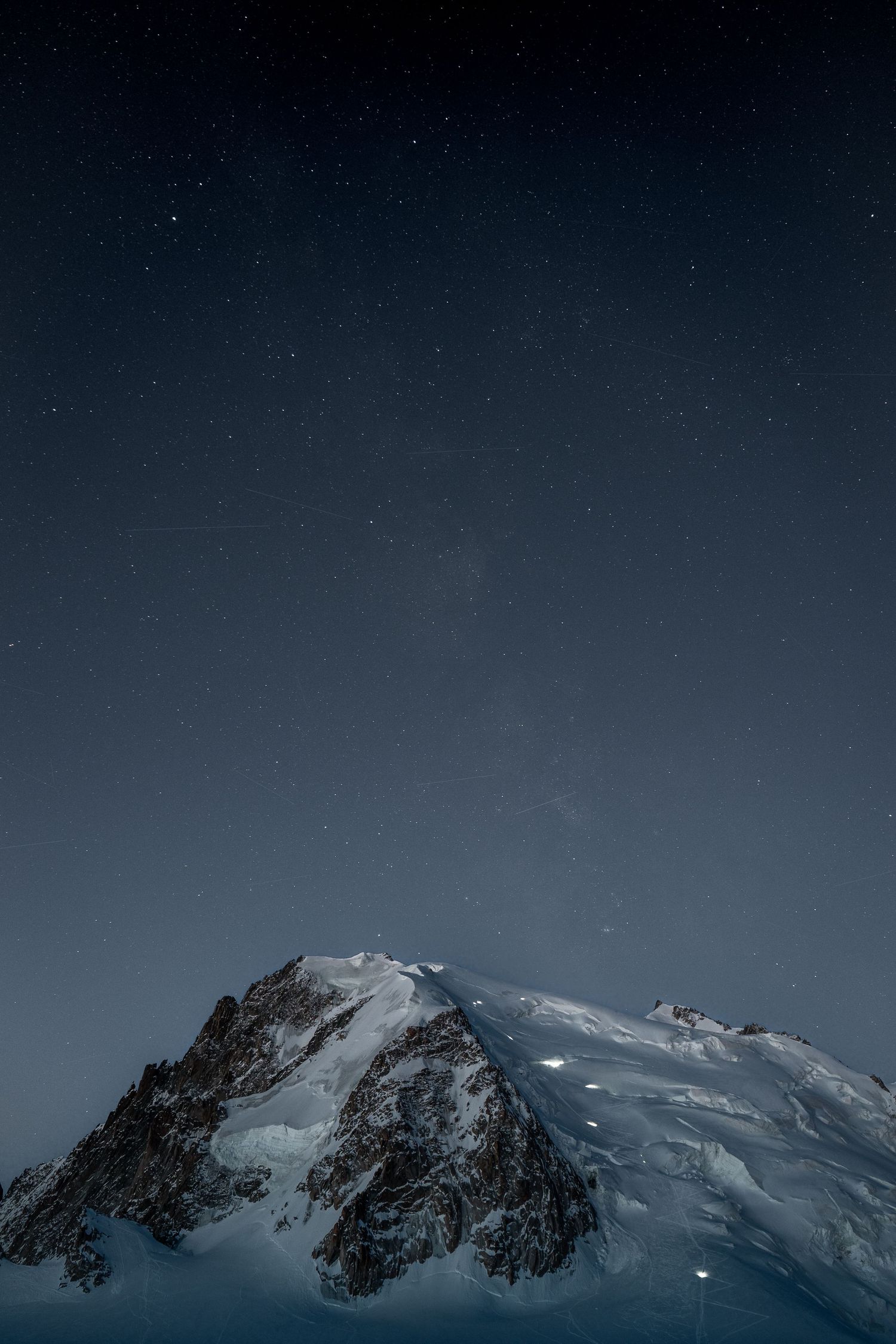Rocky snow-capped mountain peak under a clear dark blue starry night sky with visible Milky Way.