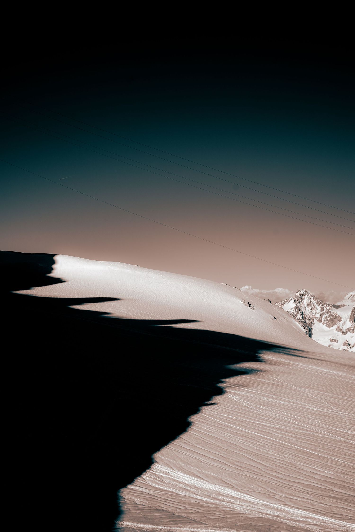 Minimalist aerial view of a snow-covered mountain ridge with stark shadows and distant peaks under a gradient sky.