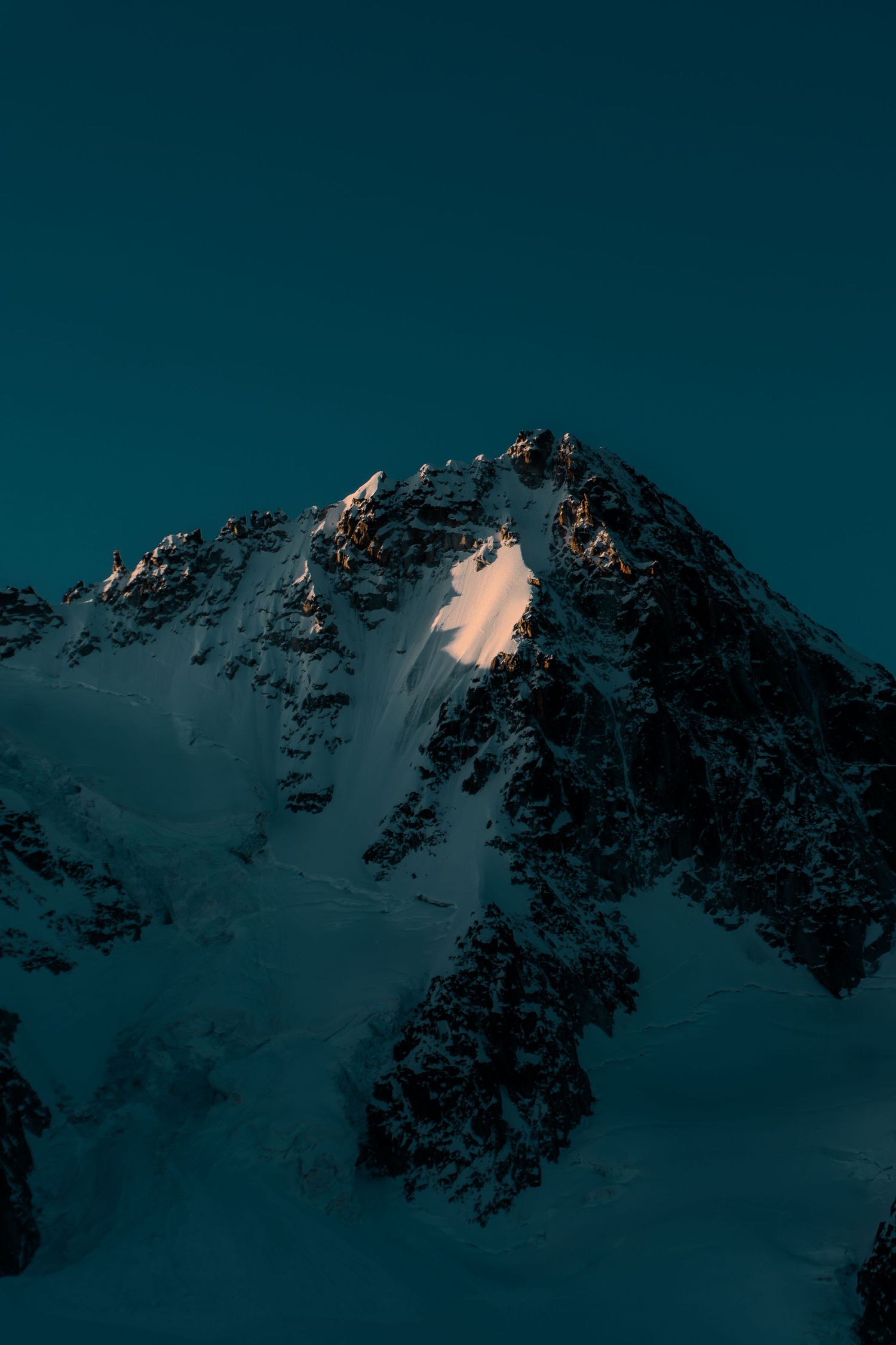 Snow-capped mountain peak illuminated by golden sunlight against a deep teal sky at dusk.