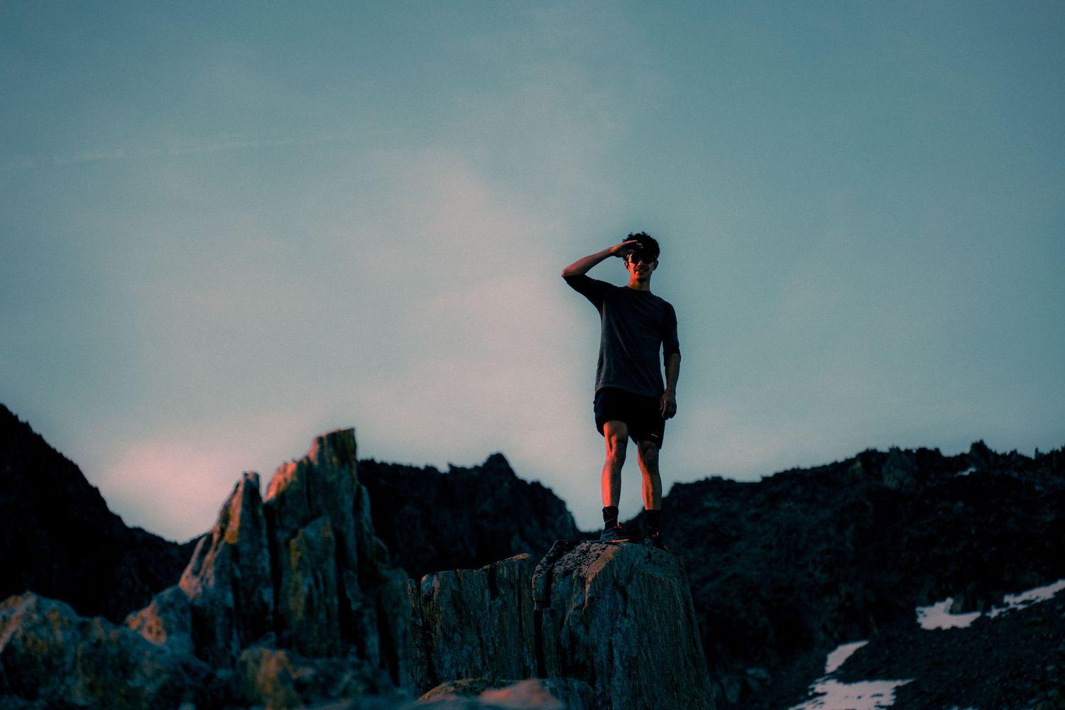 Lone hiker standing on rocky summit shielding eyes while gazing at twilight mountain landscape.