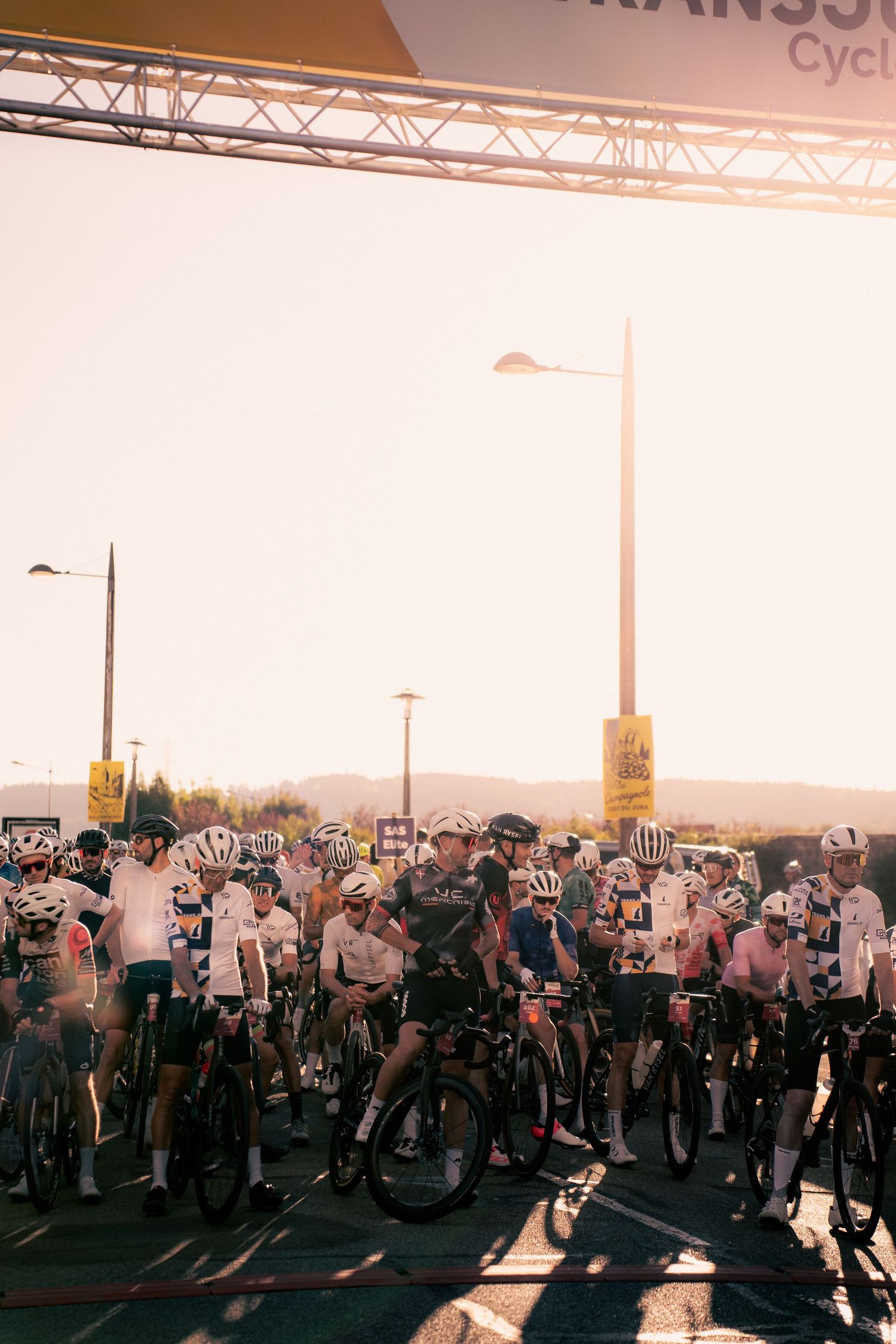 Cyclists gather at the start line of a TransSavoie cycling race event in bright sunlight.