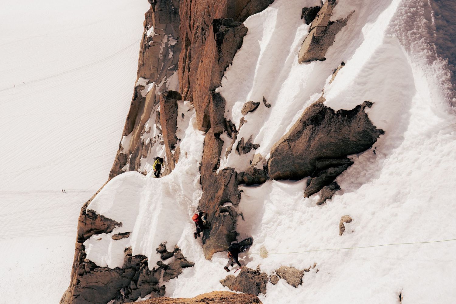 Climbers ascending a steep snow-covered rocky mountain face with dramatic cliffs and overhanging snow formations.