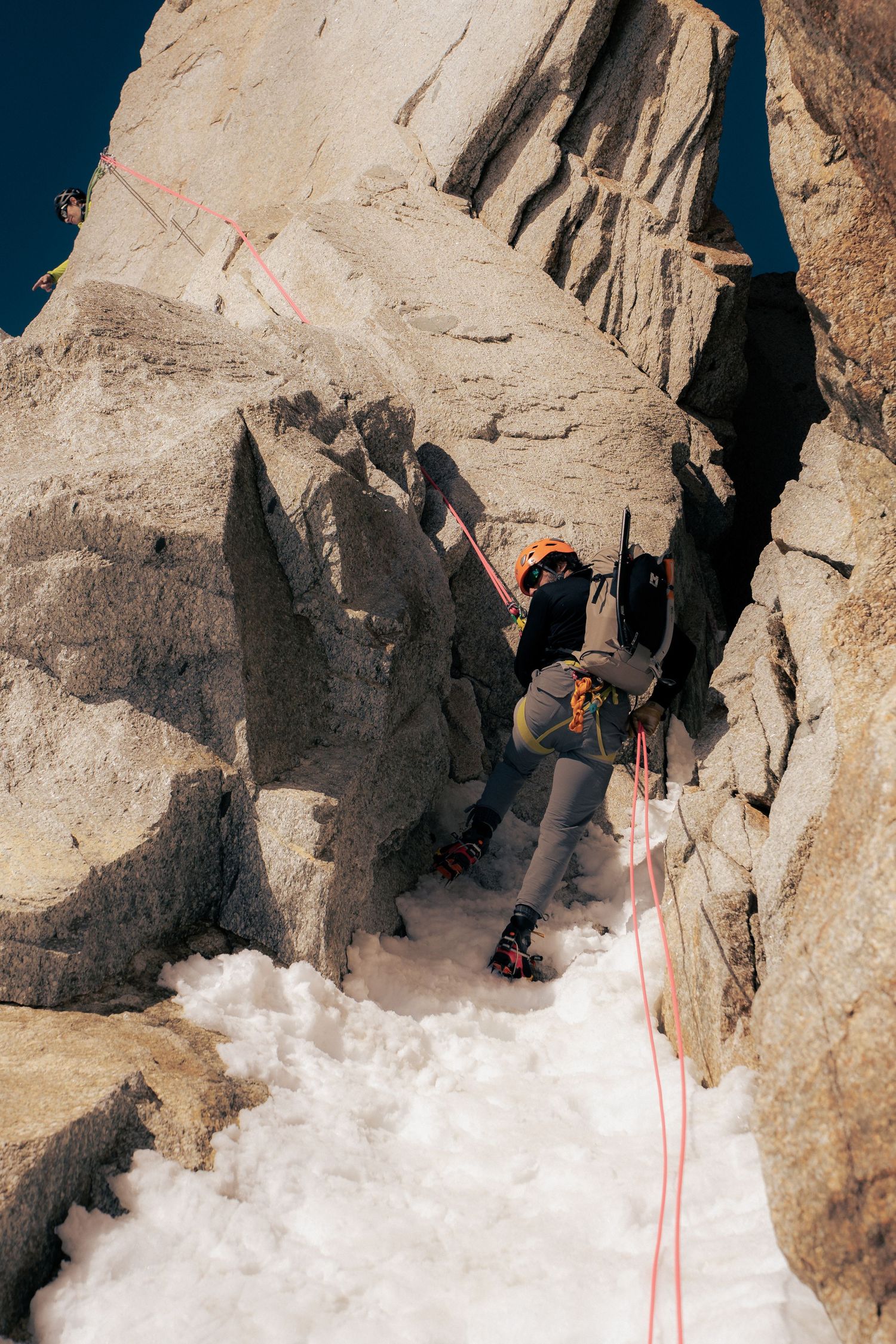 Rock climber in orange helmet ascending snowy granite crevice with red rope on a sunny alpine climb.