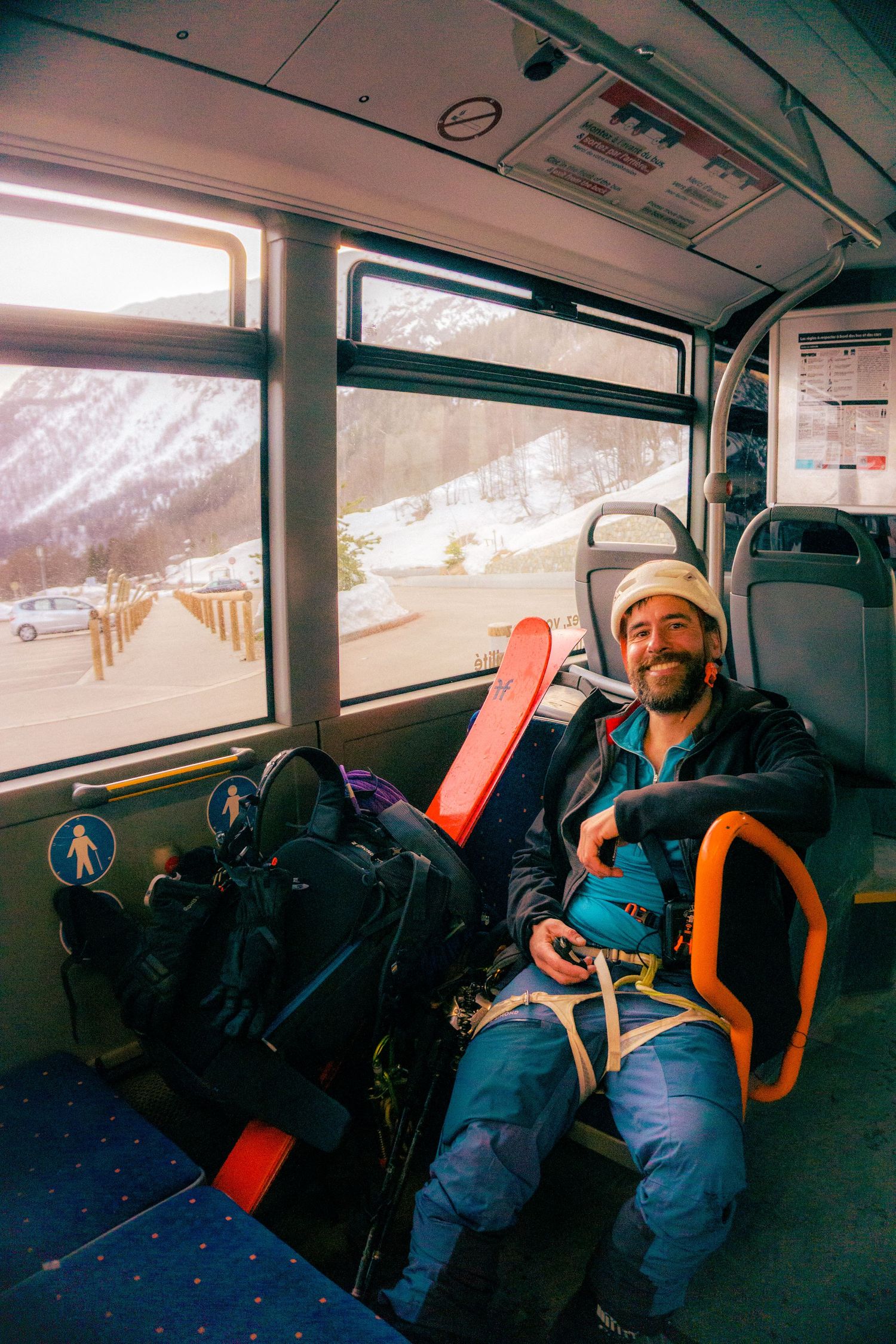 Smiling skier relaxing on a bus with ski equipment, wearing winter gear, snowy mountain landscape visible through window.