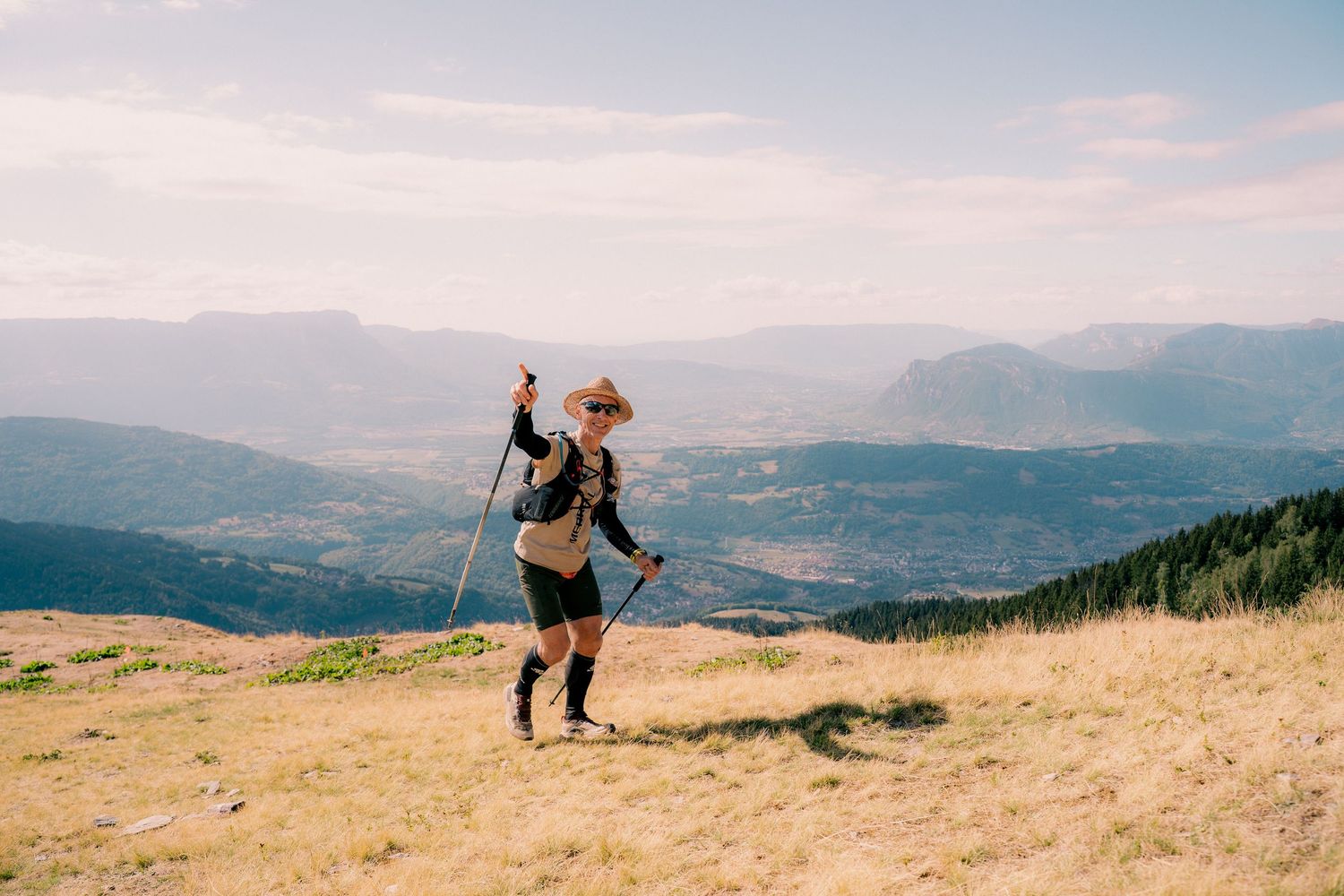 Solo hiker with trekking pole standing on open mountain summit with panoramic valley views below.