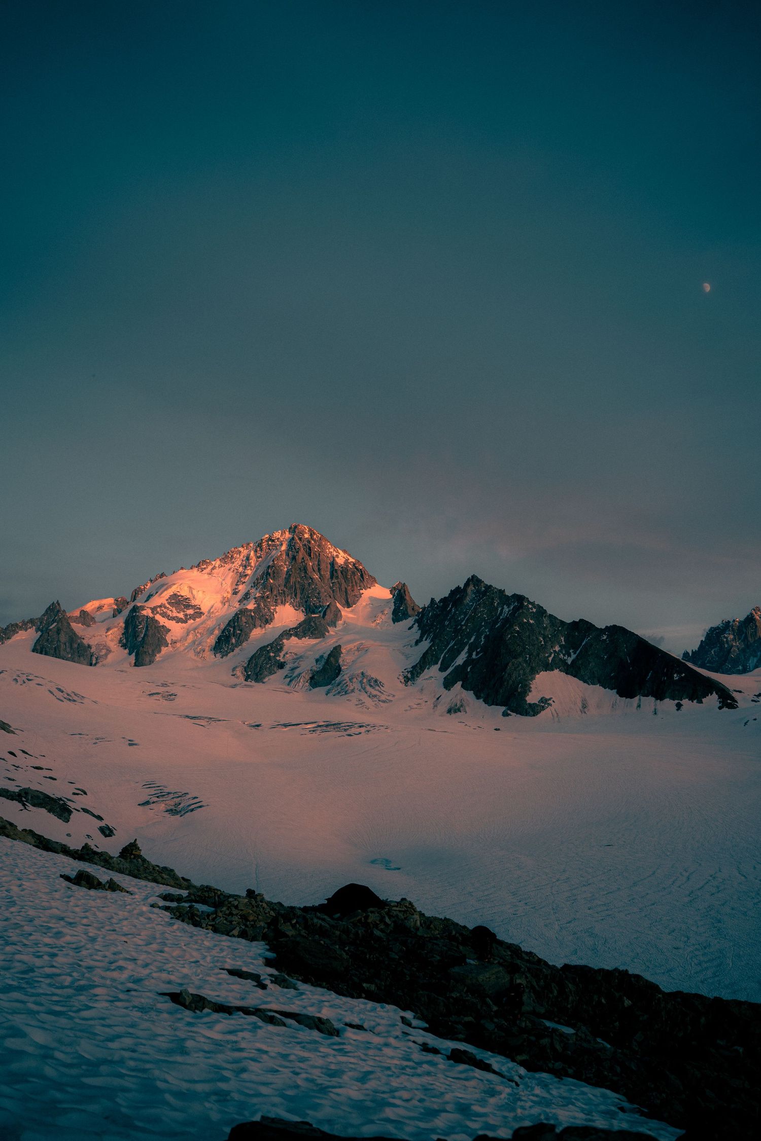 Snow-covered alpine peak glowing red at sunset with crescent moon visible in dark blue sky.