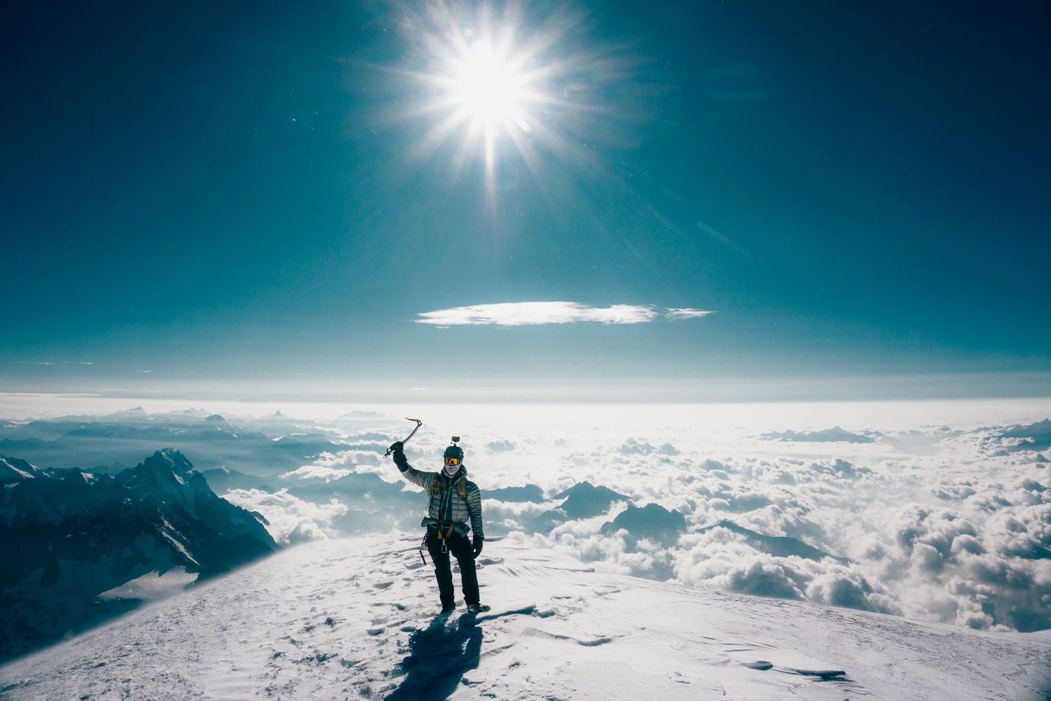 Mountain climber stands triumphant on snowy summit holding ice axe aloft under bright sun above clouds.