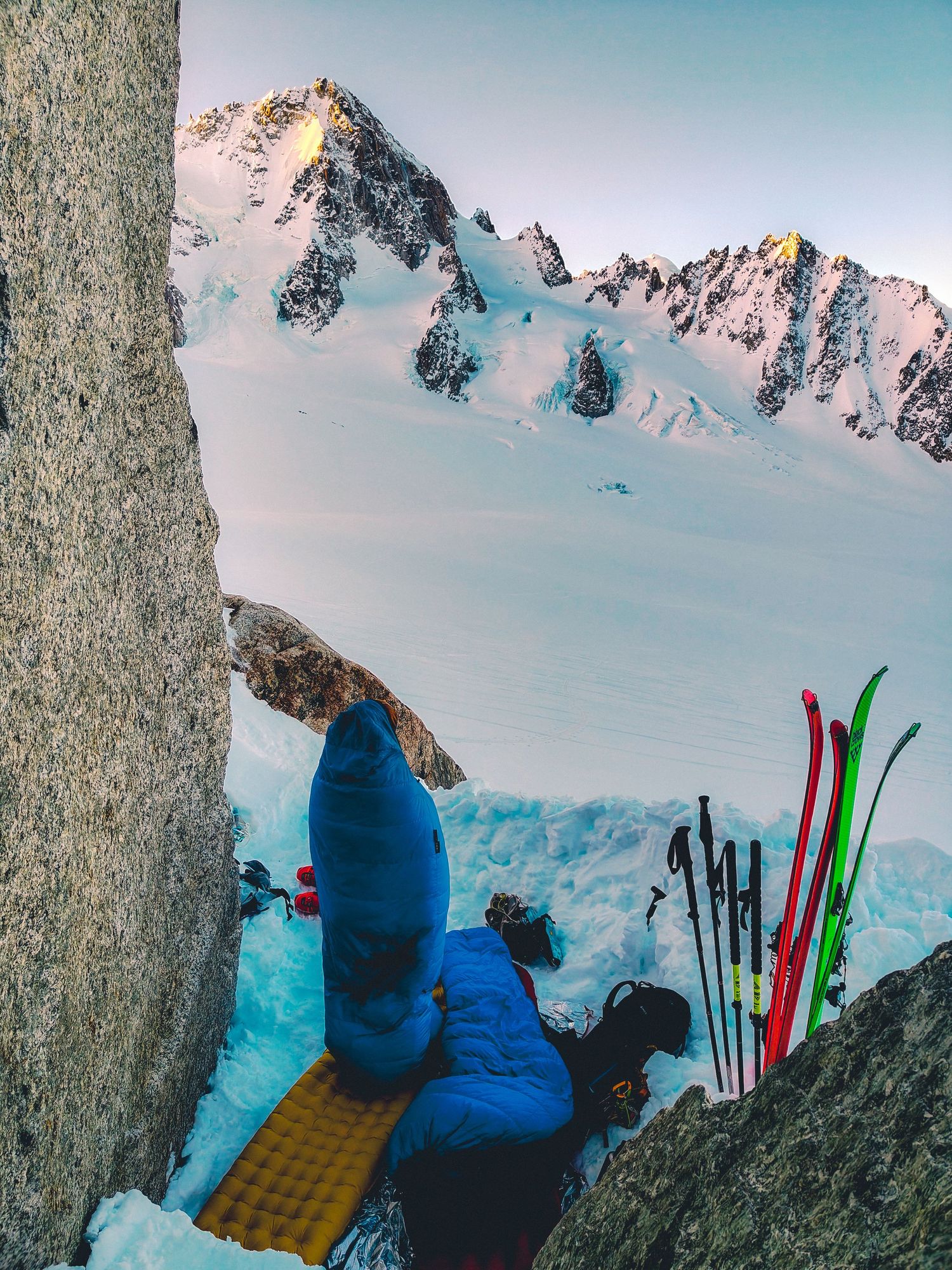 Skier camping on snowy mountain ledge with skis, poles, and sleeping pad beside dramatic alpine peaks at sunset.