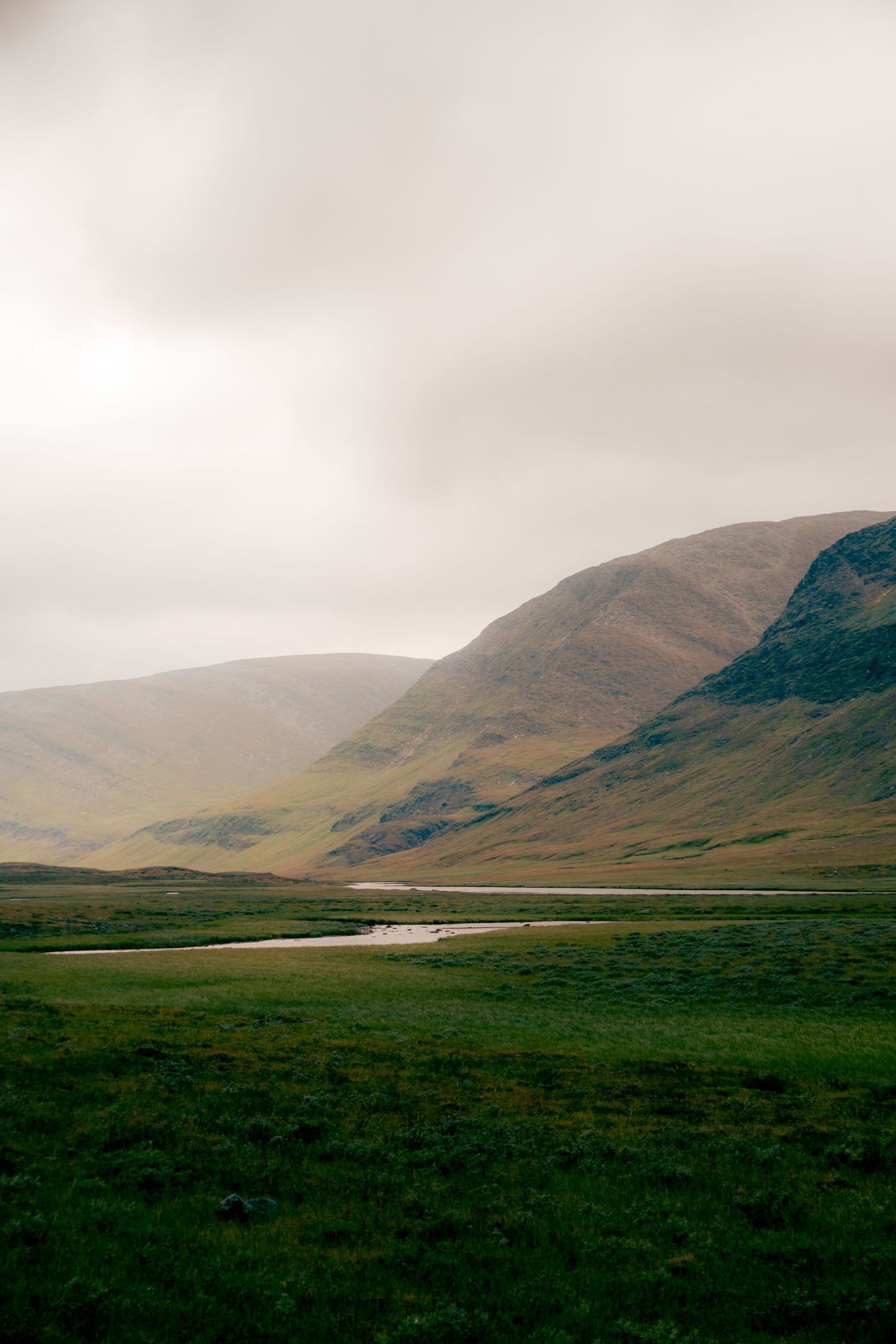 Moody landscape of green valley with winding stream and rolling hills under overcast sky in Scotland.