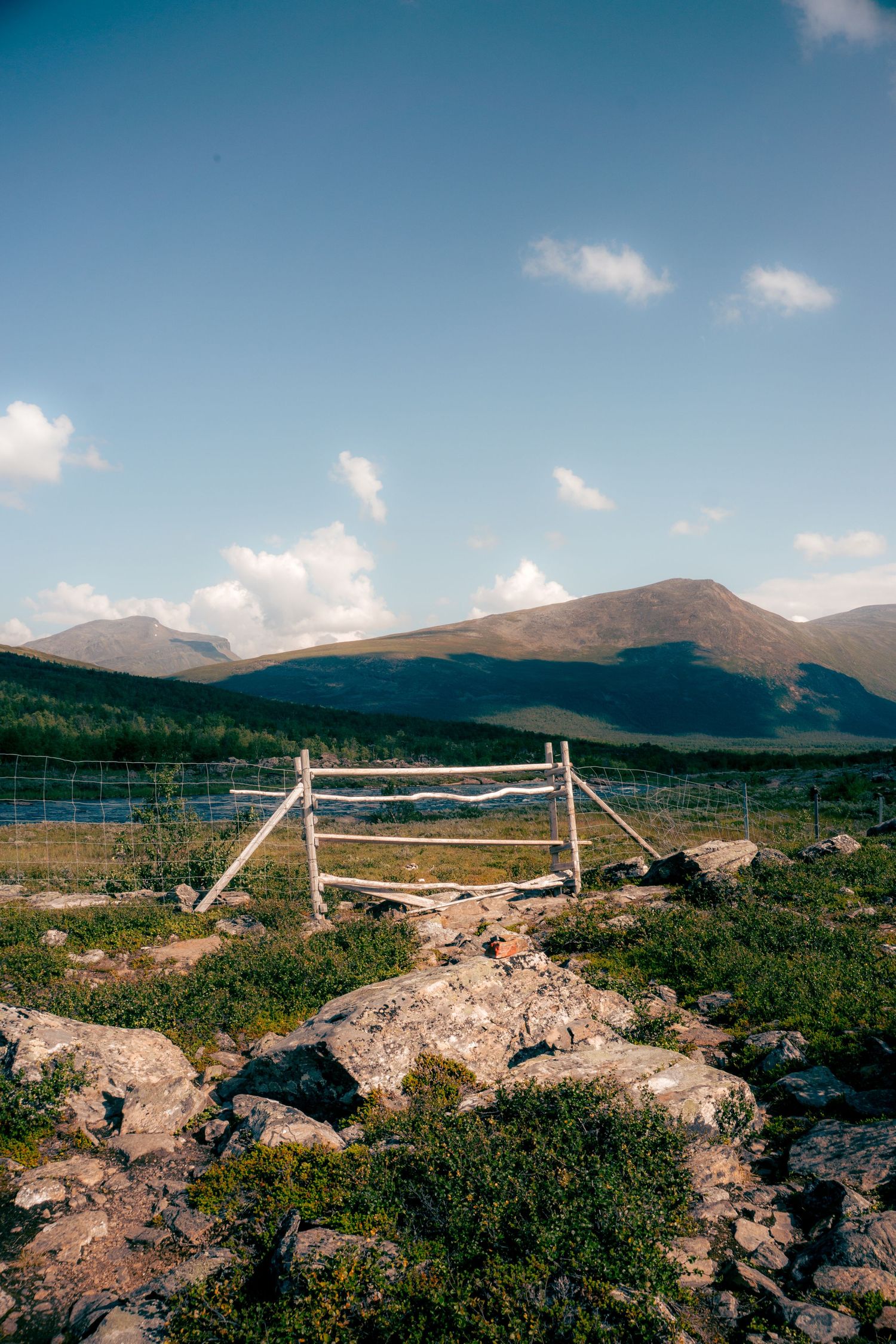 Rustic wooden gate and fence in a rocky alpine meadow with mountains and blue sky beyond.