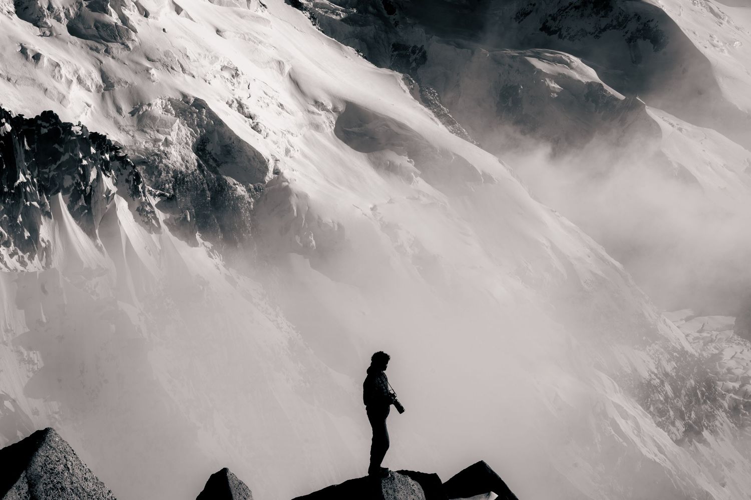 Silhouette of a lone hiker standing on rocks against dramatic snow-capped mountain peaks shrouded in mist.