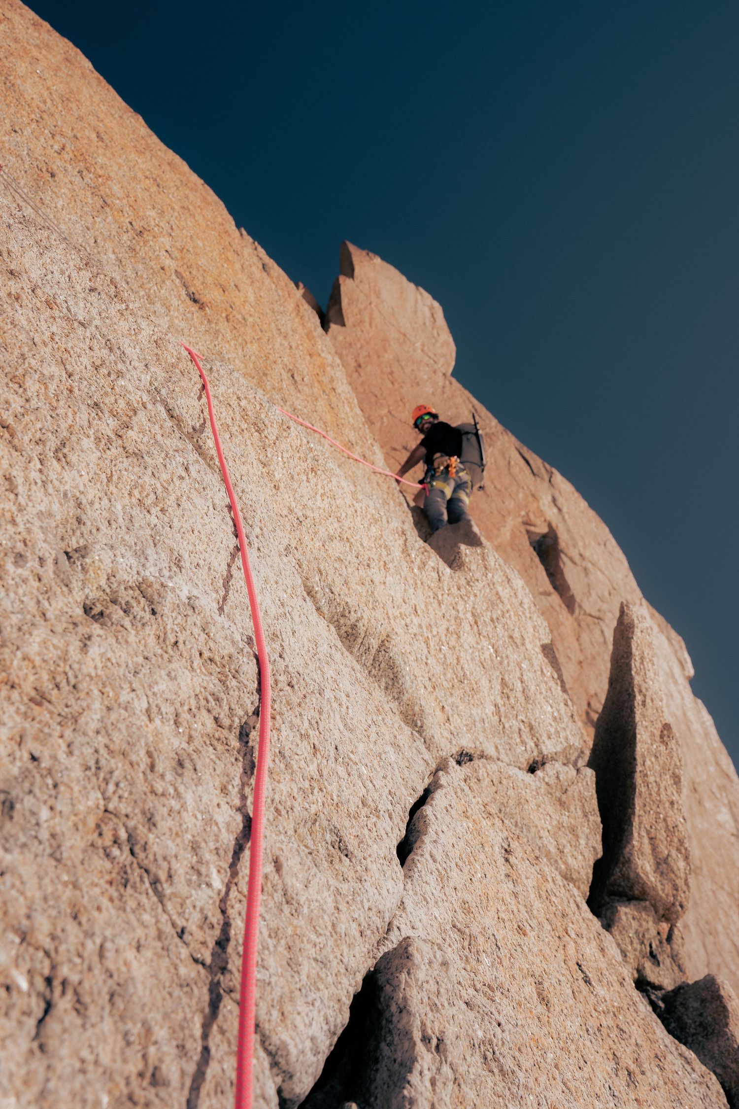 Rock climber scaling a steep granite face with a red rope, against a clear blue sky.