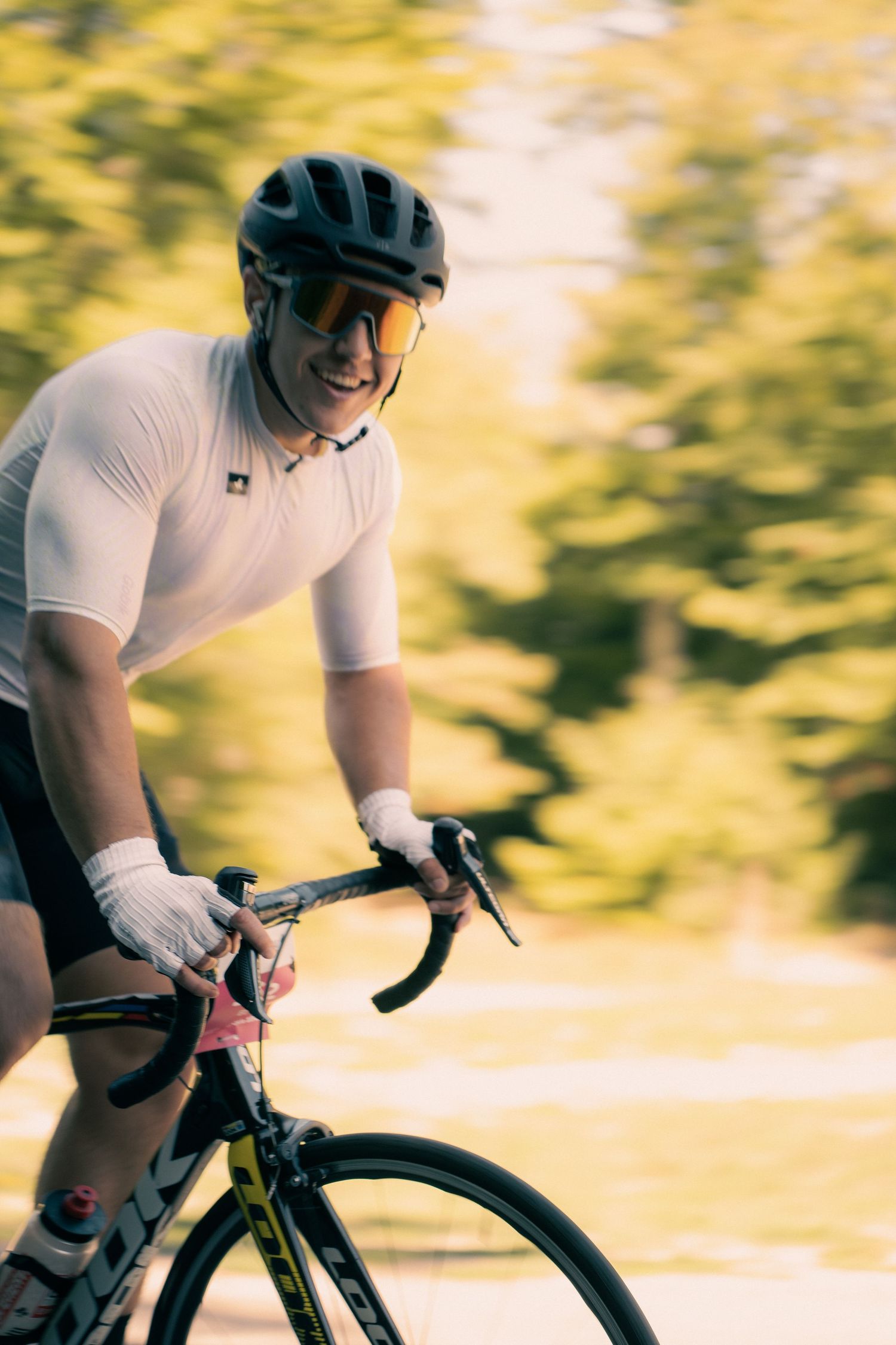 Smiling male cyclist in white jersey and helmet rides road bike at speed with blurred green trees in background.