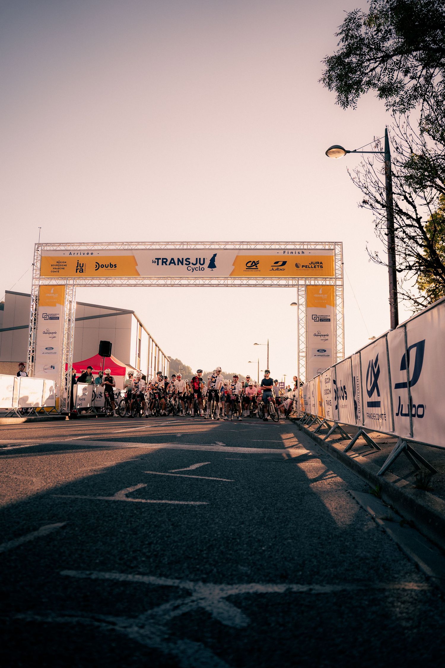 Cyclists gather at the start line of the TransJu cycling race, with sponsor banners overhead on a sunny day.