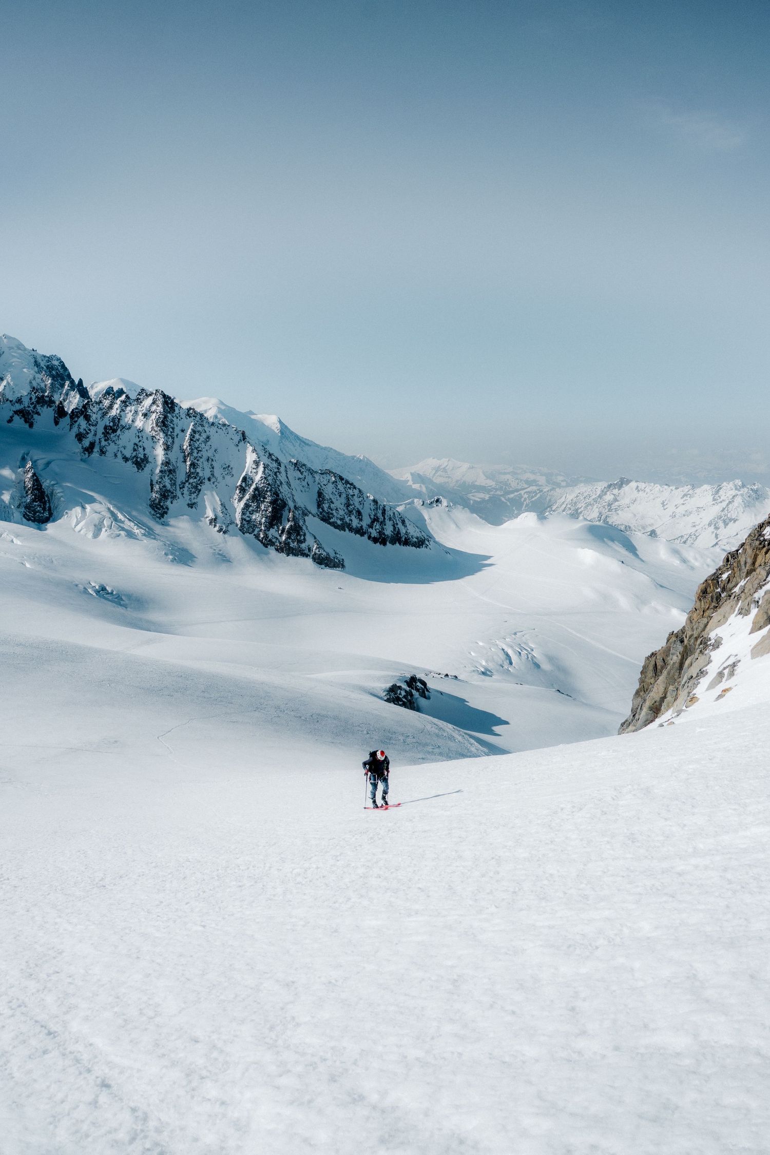 A lone hiker traverses a vast snowy alpine landscape surrounded by snow-capped mountain peaks under a clear blue sky.