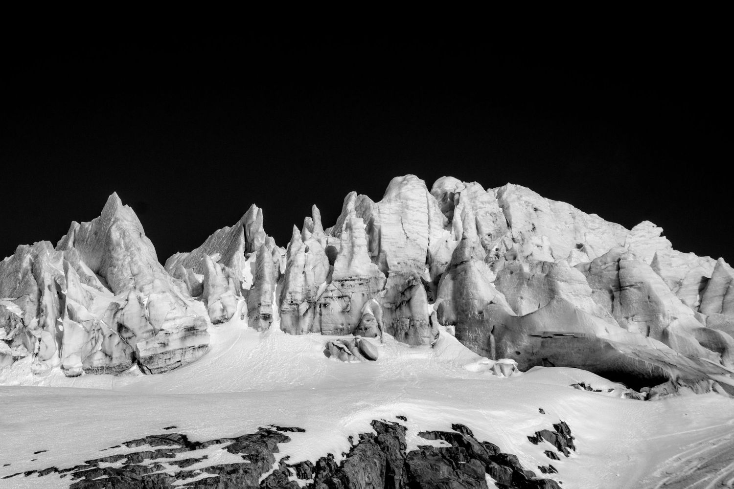 Black and white photo of jagged glacier ice formations rising against a dark sky in a snowy Arctic landscape.