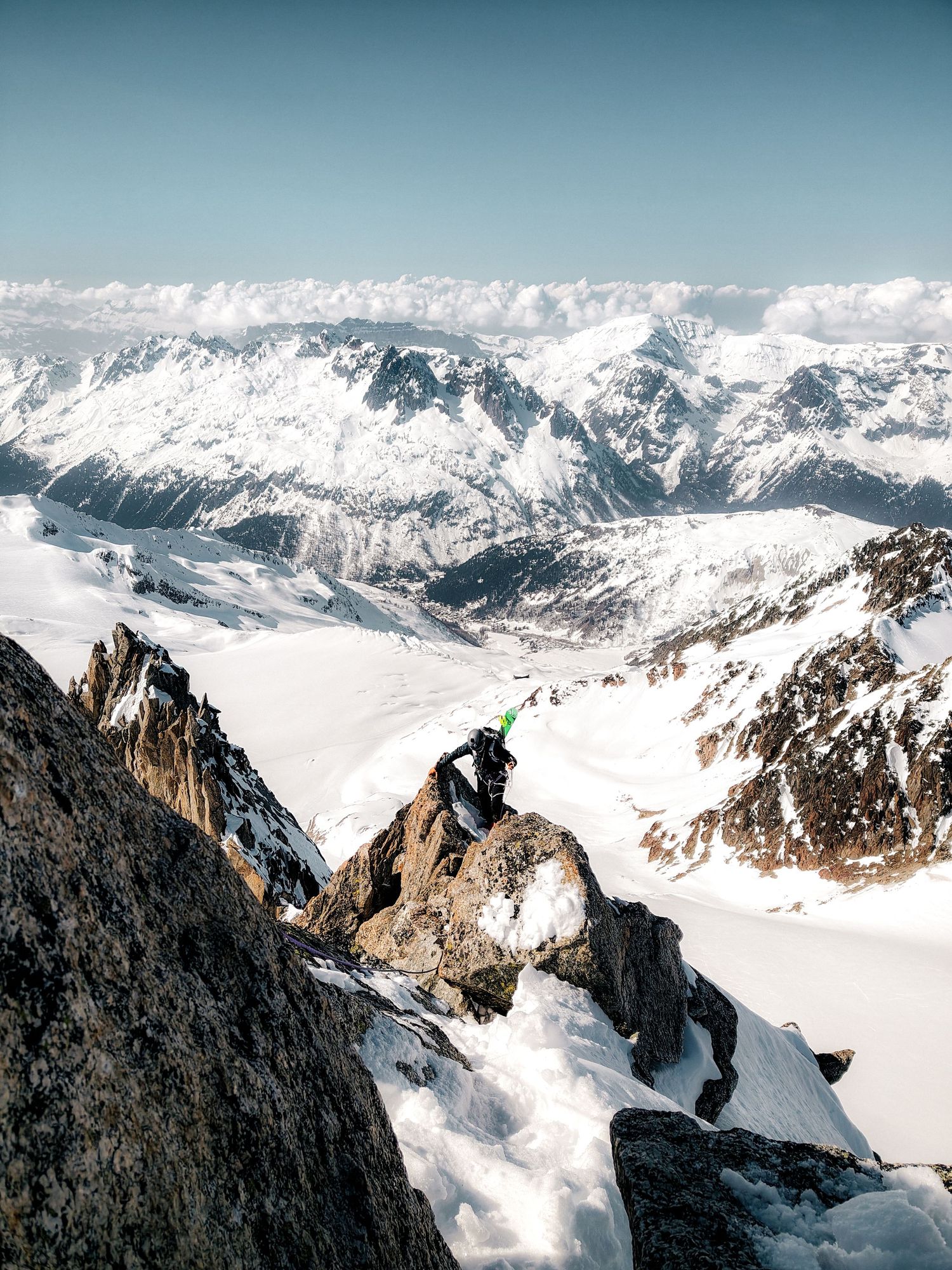 A mountaineer perched on rocky summit with vast snow-covered Alpine peaks and glaciers stretching into the horizon below.