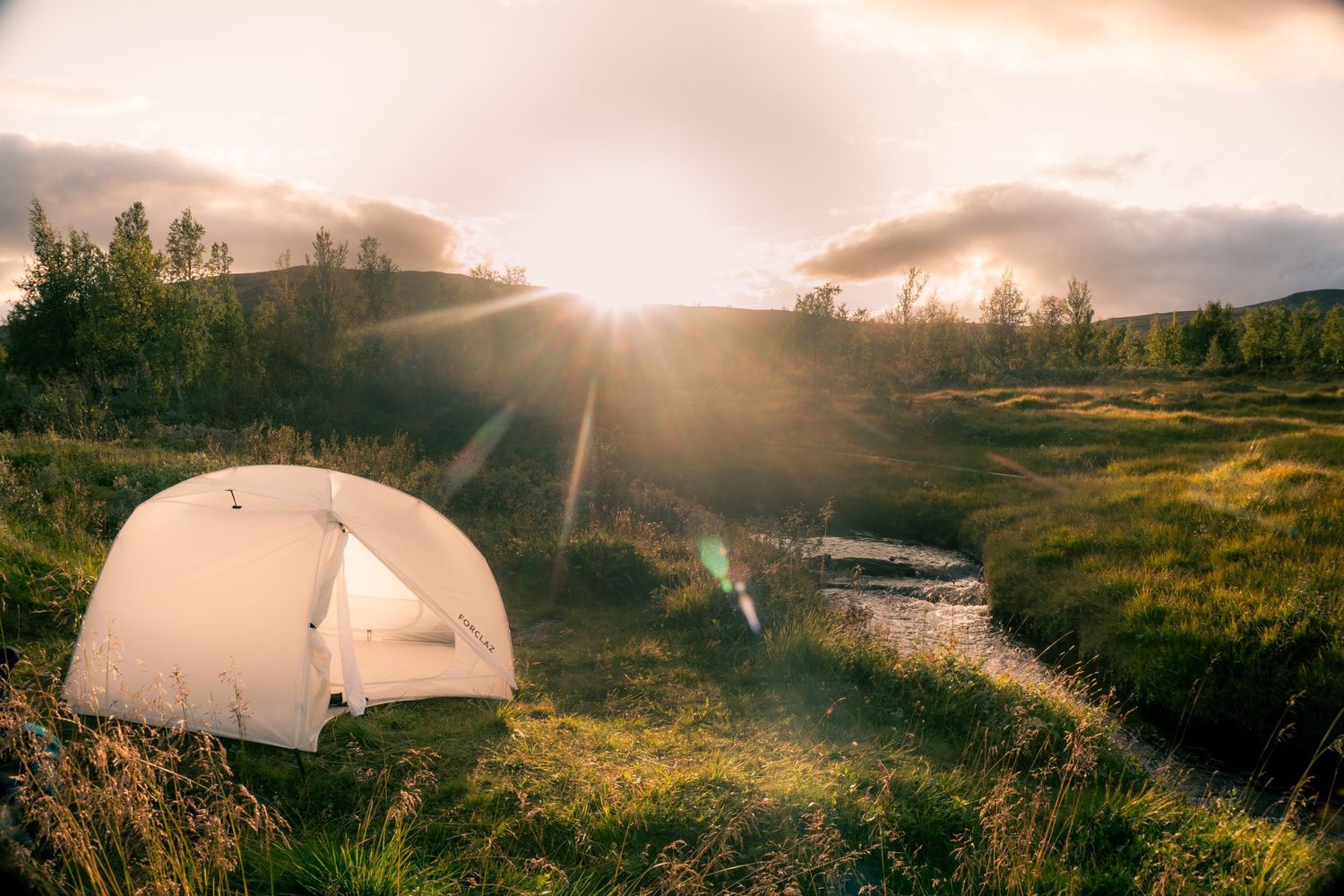 A white camping tent glows in golden sunset light beside a stream in a green valley.
