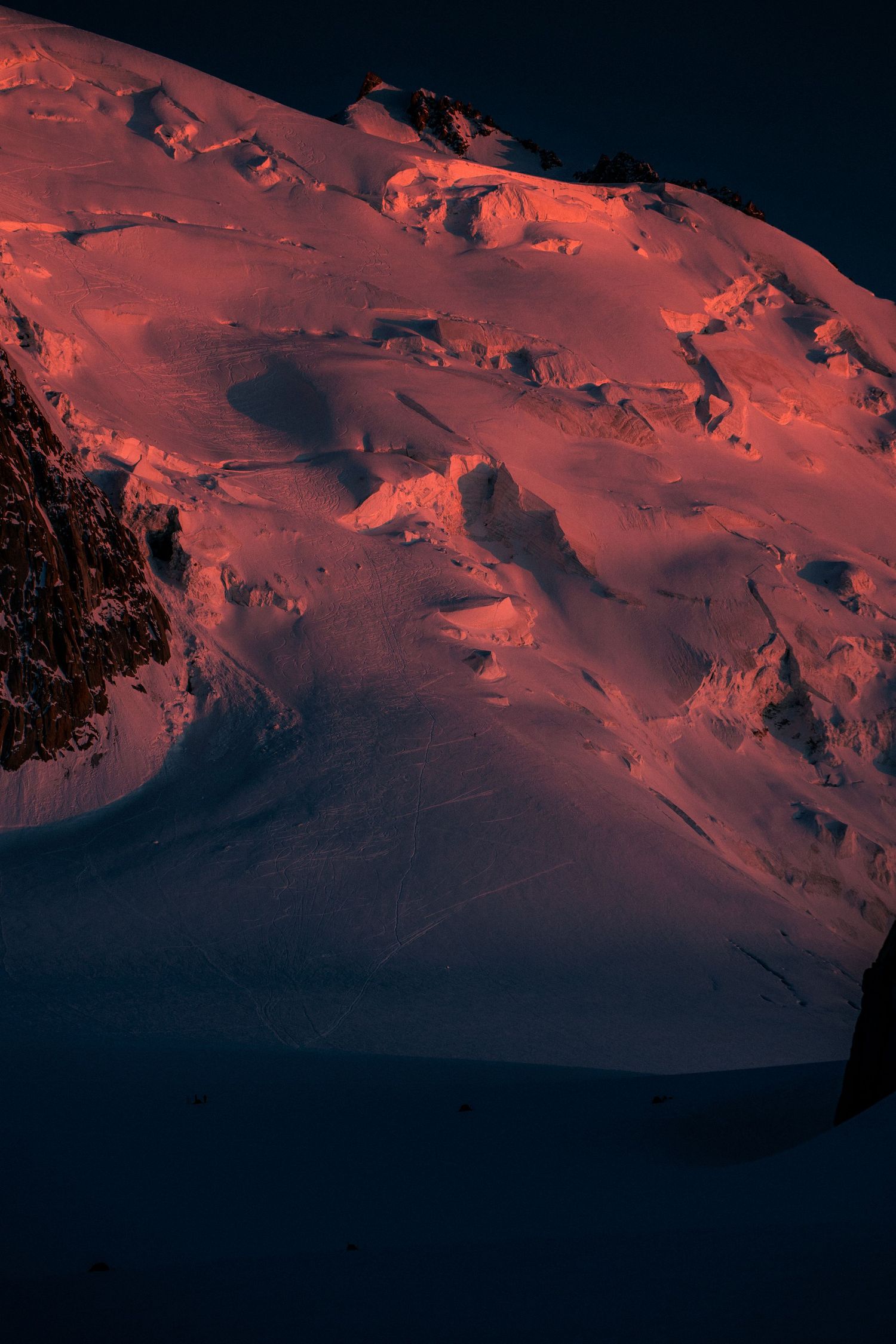 Close-up of a steep snow and ice mountain face glowing deep orange-red in alpenglow at sunset.