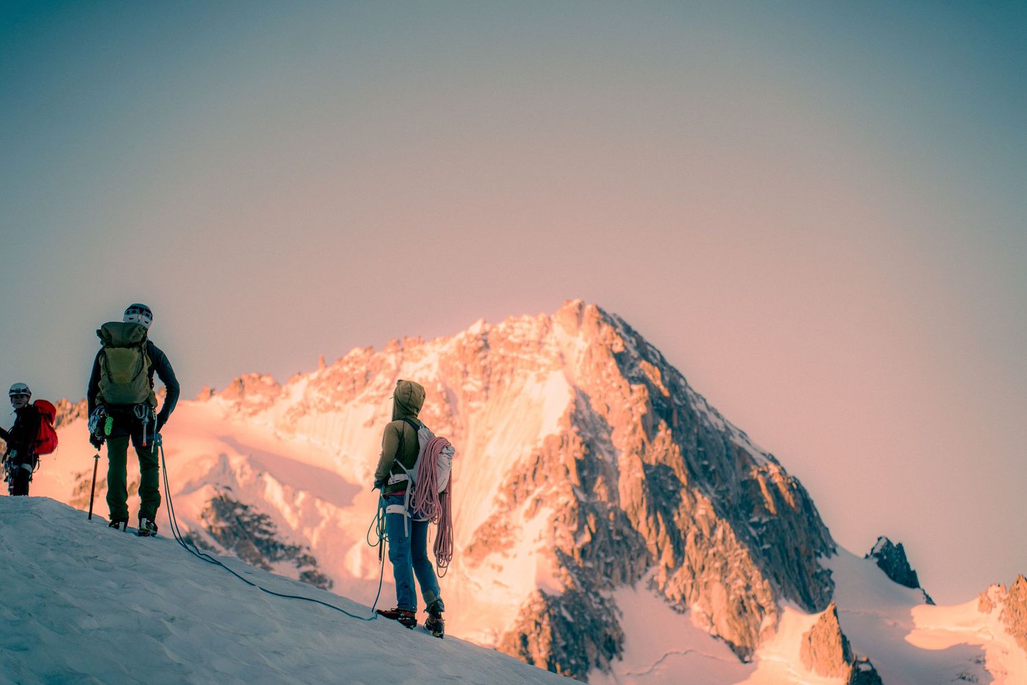 Alpine climbers ascending snowy ridge with towering snow-capped mountain peak glowing in golden sunrise light.