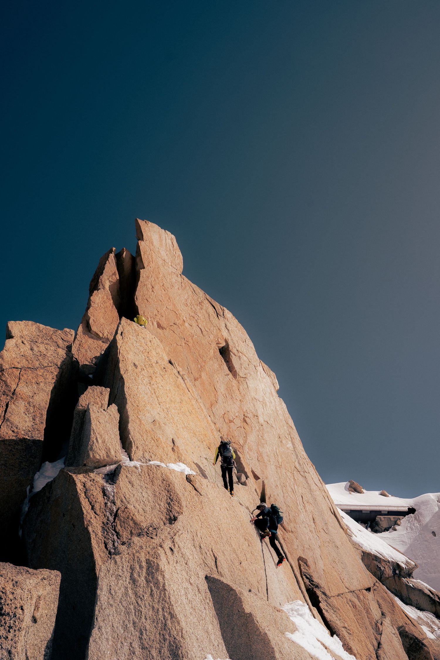 Two climbers ascending a jagged rocky mountain face with snow patches under a clear deep blue sky.
