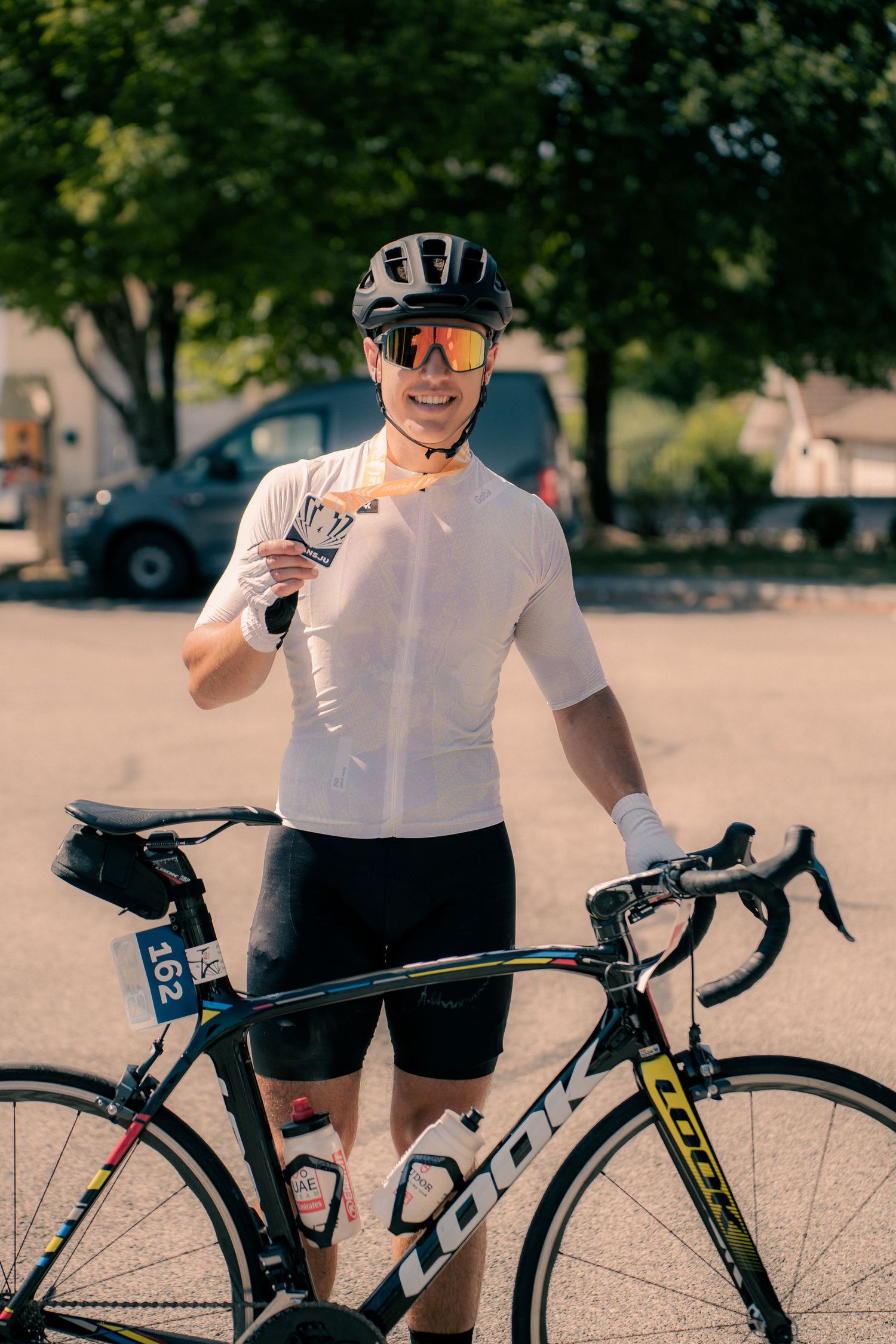 Cyclist in white jersey and helmet stands smiling beside a Look road bike in a sunny parking lot.